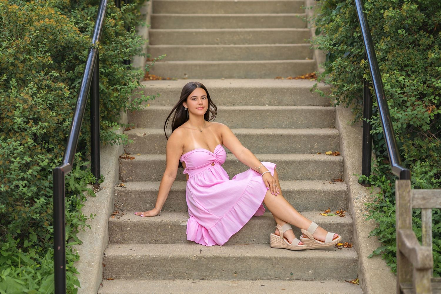 Young woman in pink sundress and wedge sandals sitting gracefully on outdoor stone steps surrounded by lush greenery.