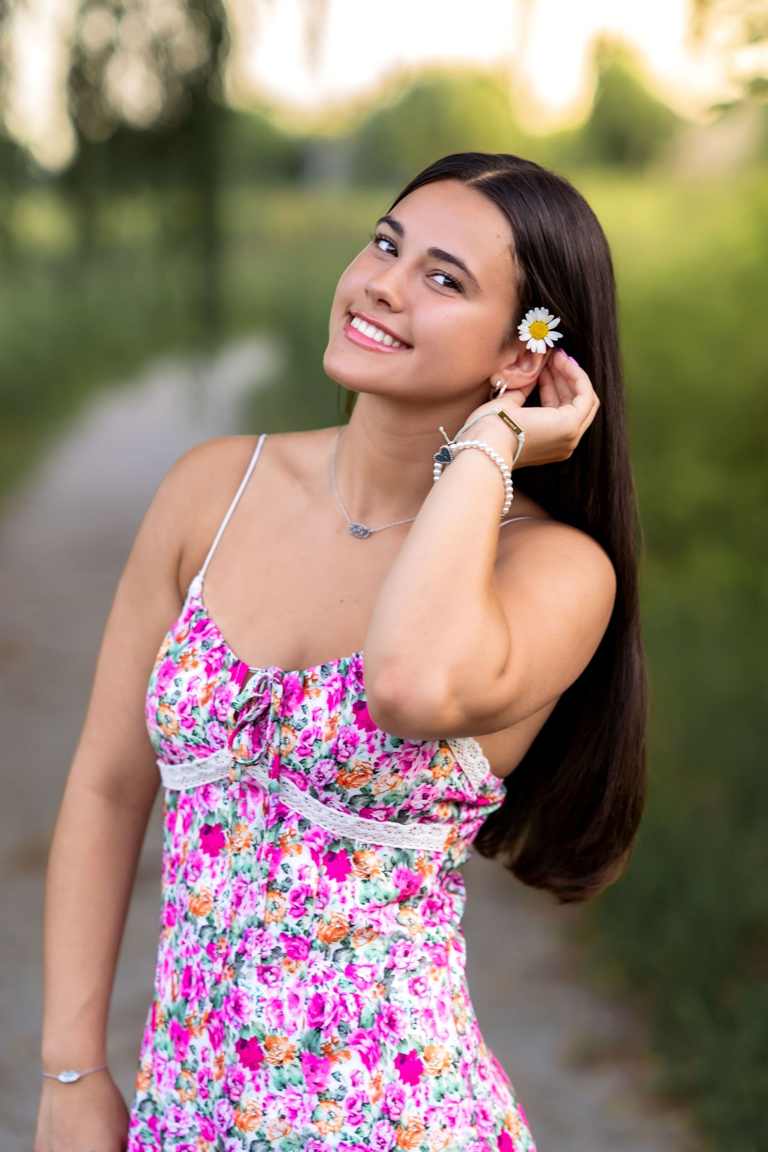 Young woman in floral sundress smiling outdoors, holding a daisy flower near her ear on a sunny day.