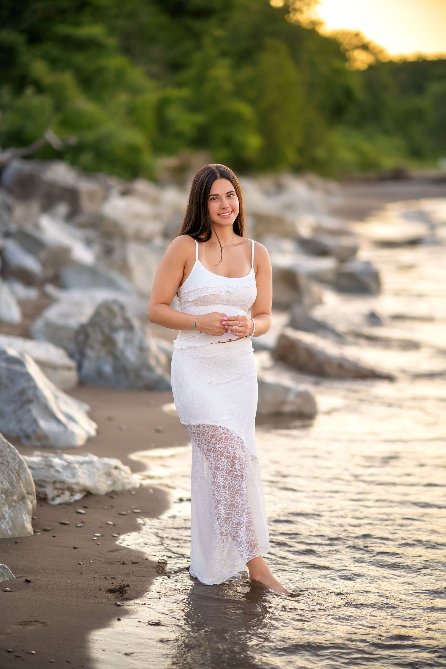 Smiling pregnant woman in white lace dress standing barefoot in shallow water on a rocky beach at sunset.