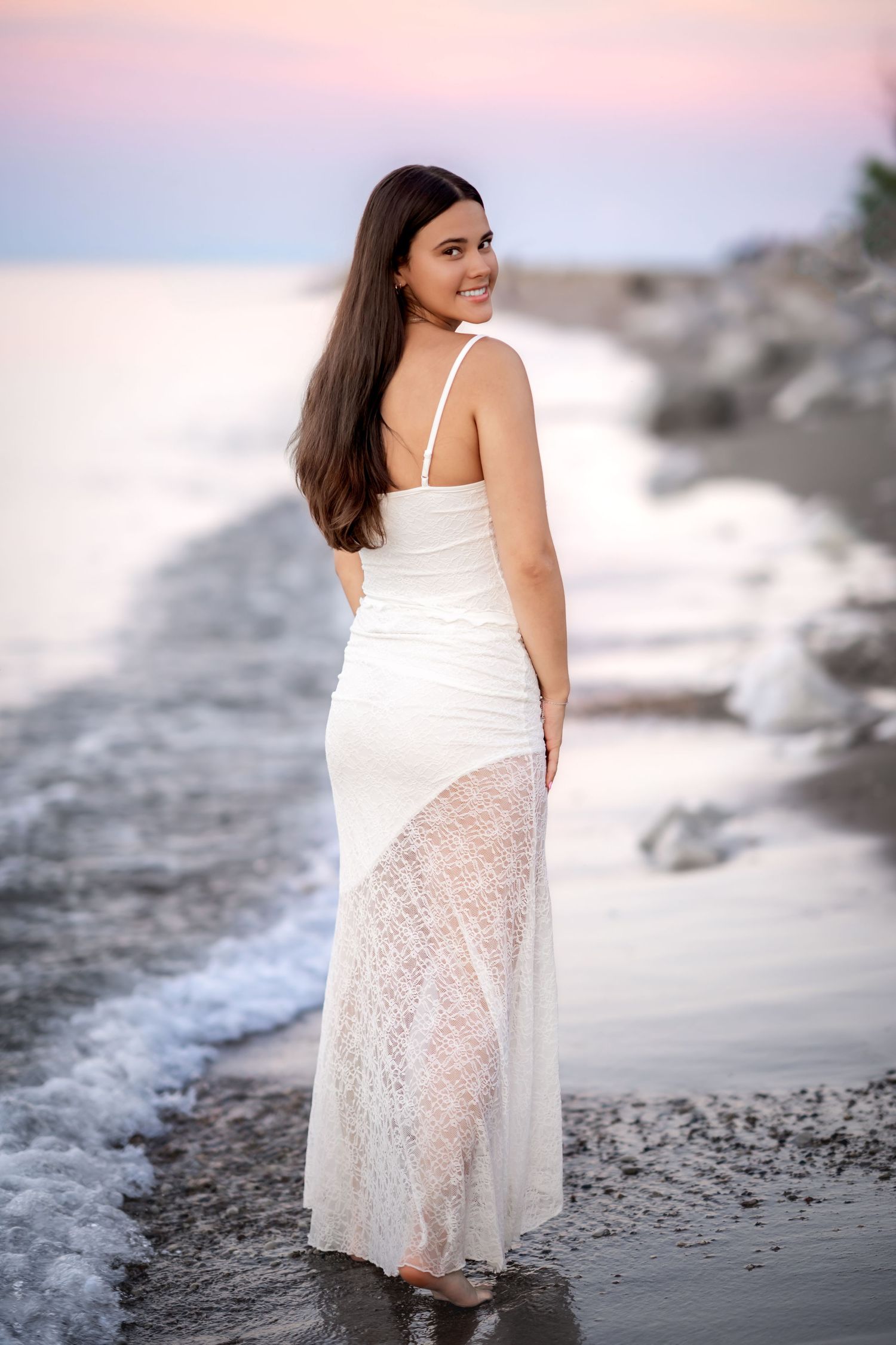 A smiling brunette woman in a white lace maxi dress stands on a rocky beach at sunset, looking back over her shoulder.