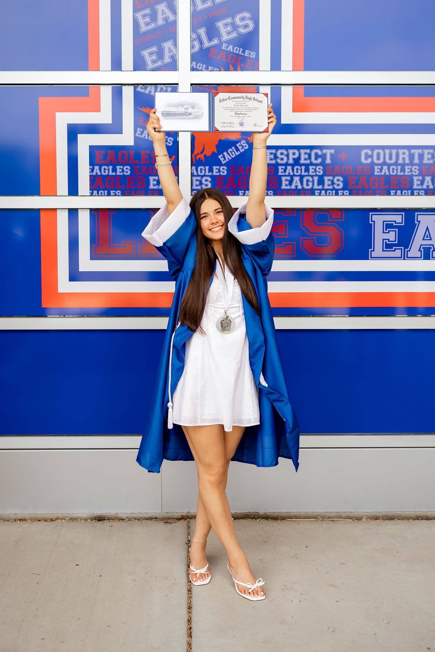 A young woman in a blue graduation gown holds diplomas overhead, posing in front of an Eagles school wall.