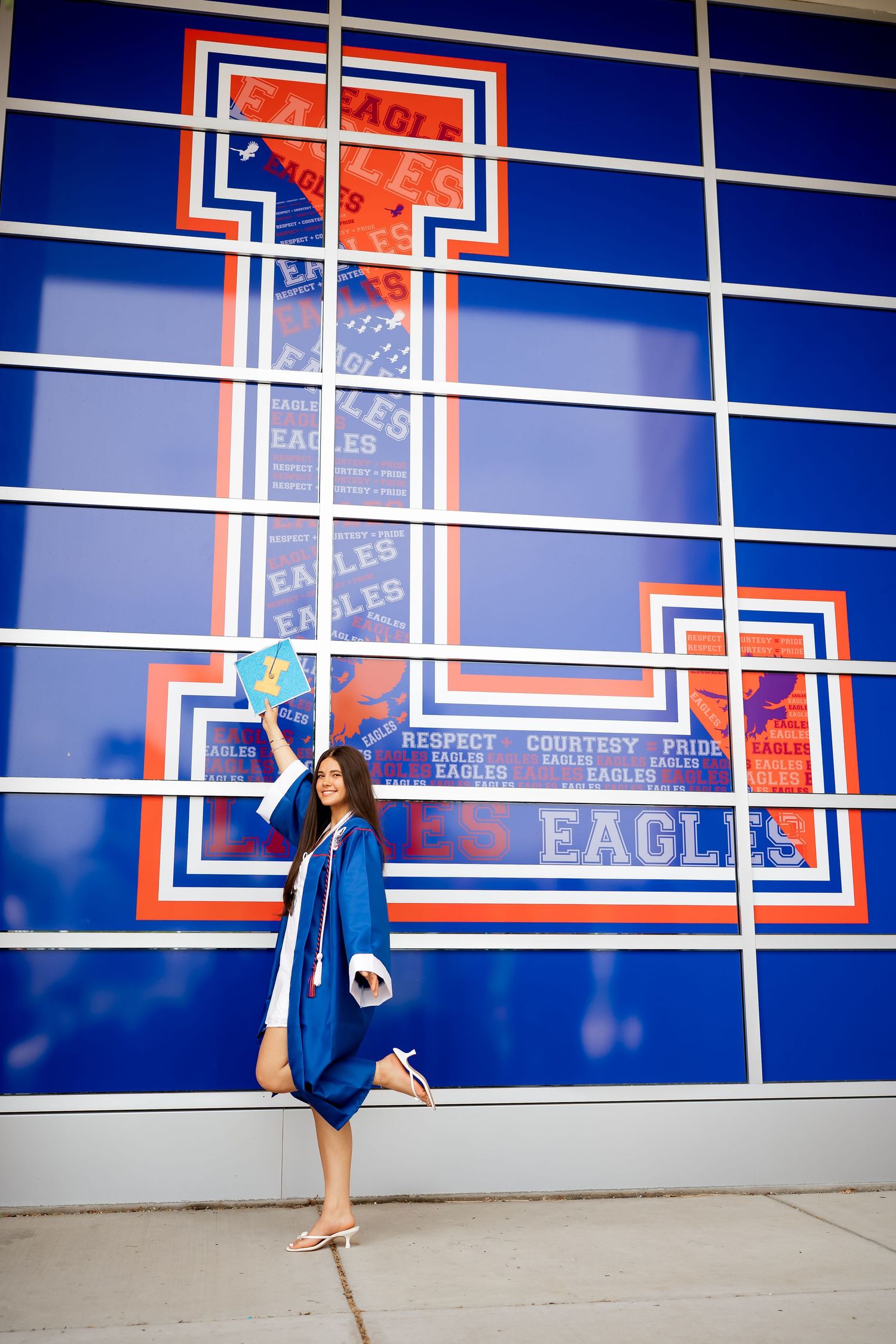 Graduate in blue cap and gown poses proudly in front of an Eagles school mural with bold blue and orange colors.