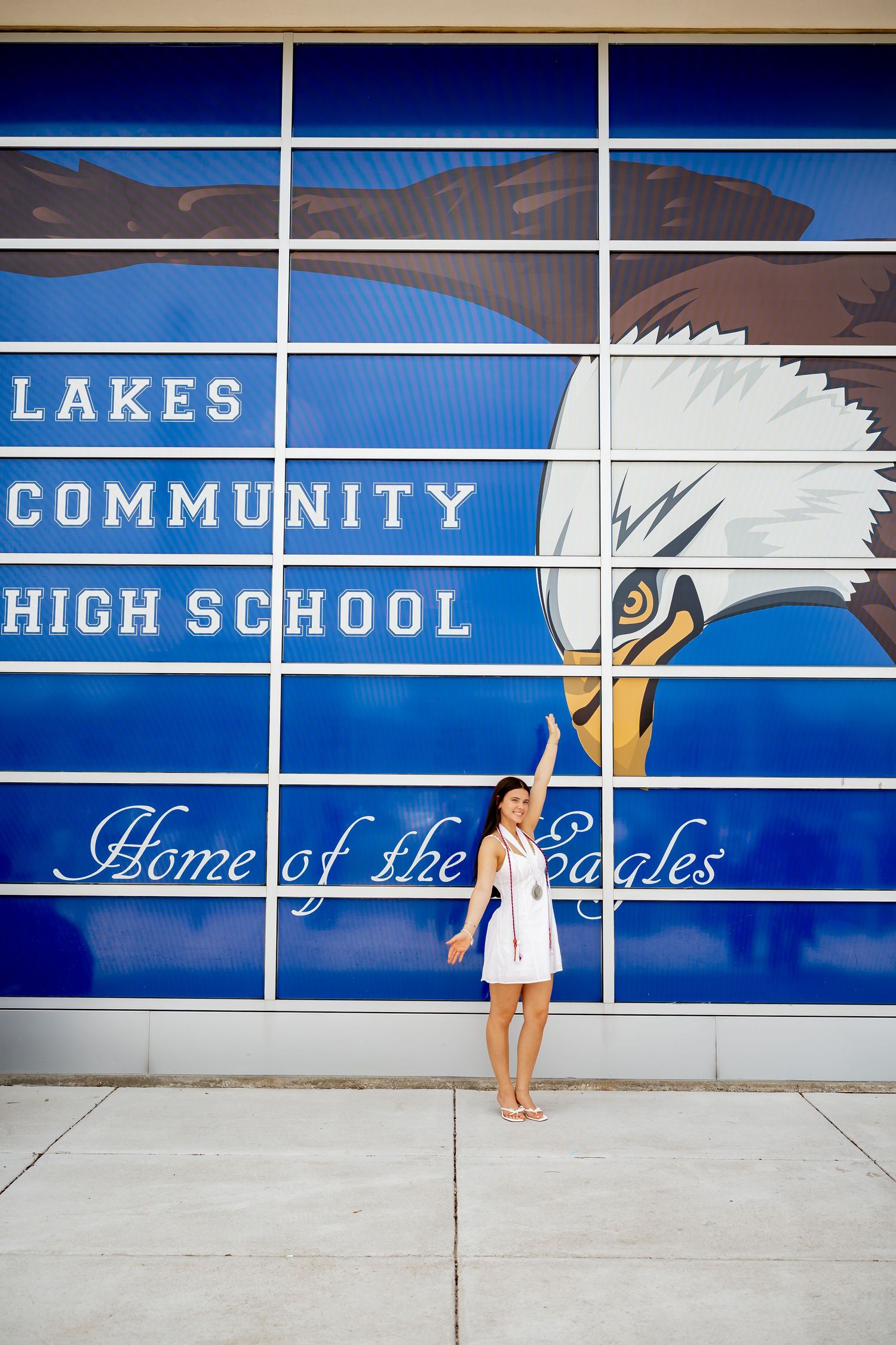 A young woman in a white dress poses in front of Lakes Community High School, Home of the Eagles mural.