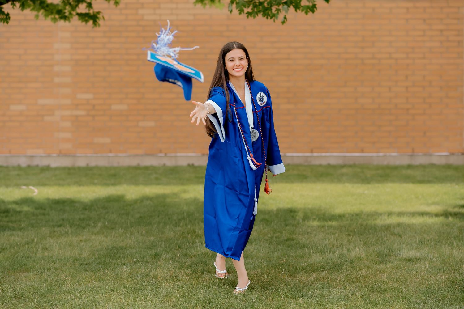 Young woman in blue graduation gown holding balloon on grass in front of brick wall.