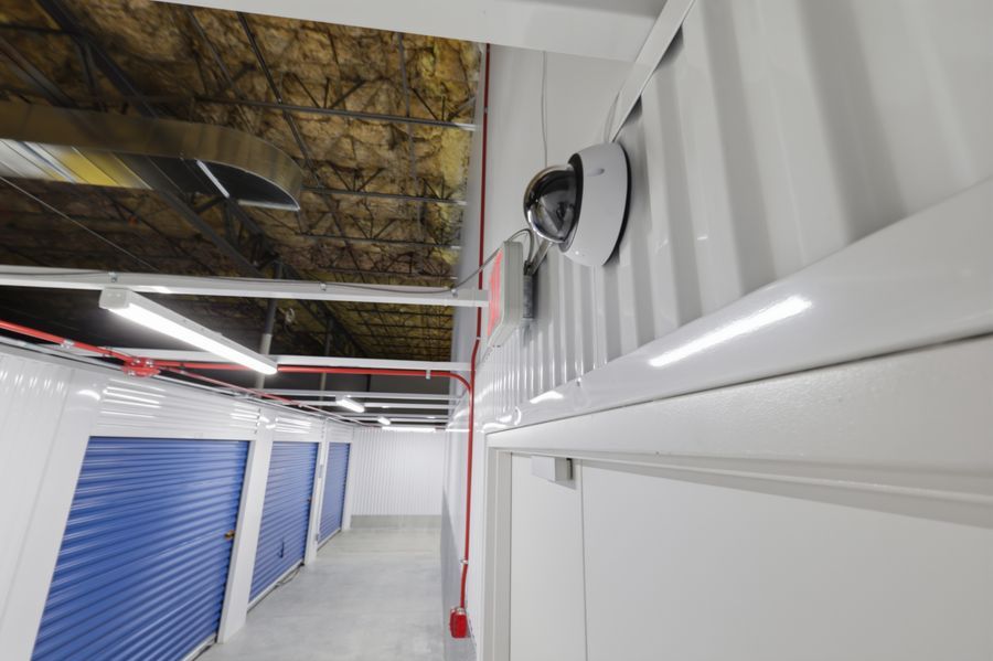 Interior storage unit corridor with blue roll-up doors, security camera, and industrial ceiling lighting.