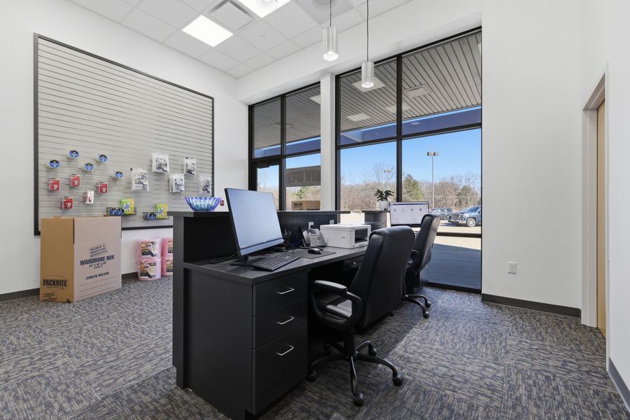 Modern self-storage facility office with black desk, computer workstations, ergonomic chairs, and large windows overlooking parking lot.