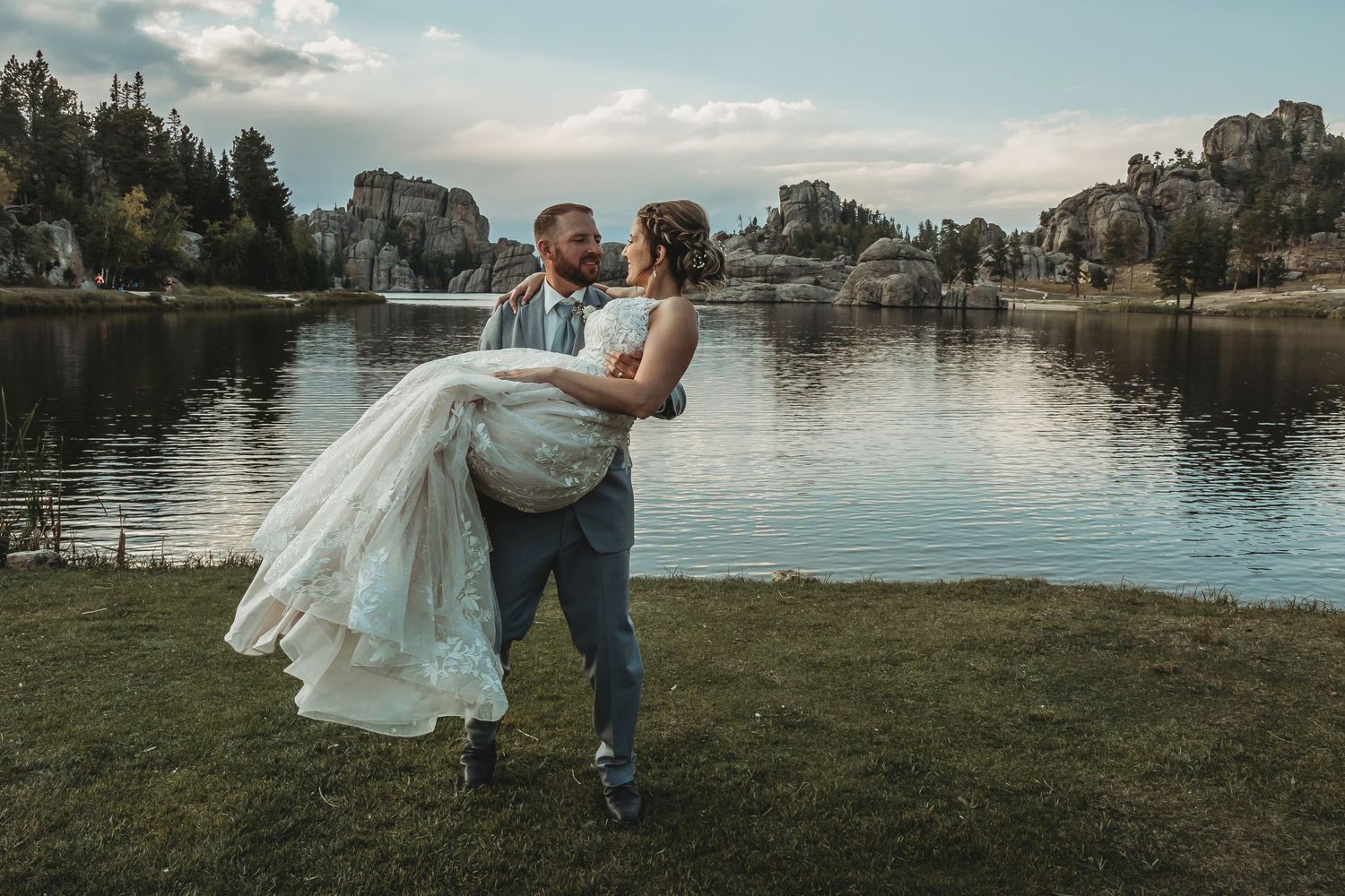 Groom carries bride in wedding dress by a scenic lake with rocky formations and trees at dusk.