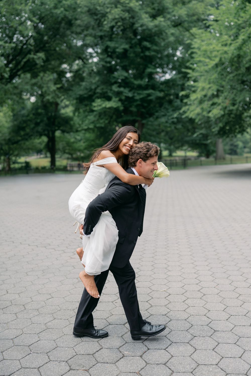Groom giving bride a piggyback ride in a park, both smiling, wearing wedding attire outdoors.