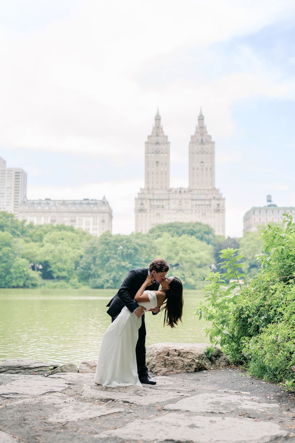 A couple shares a romantic dip kiss in Central Park with the San Remo building and lake in the background.