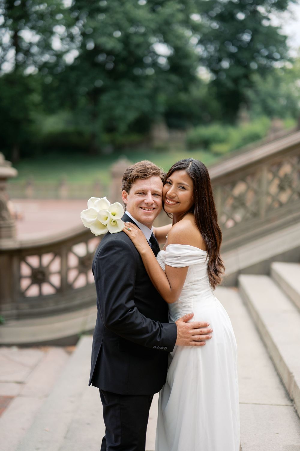 A smiling bride and groom embrace on stone steps in Central Park, NYC, with lush green trees in the background.