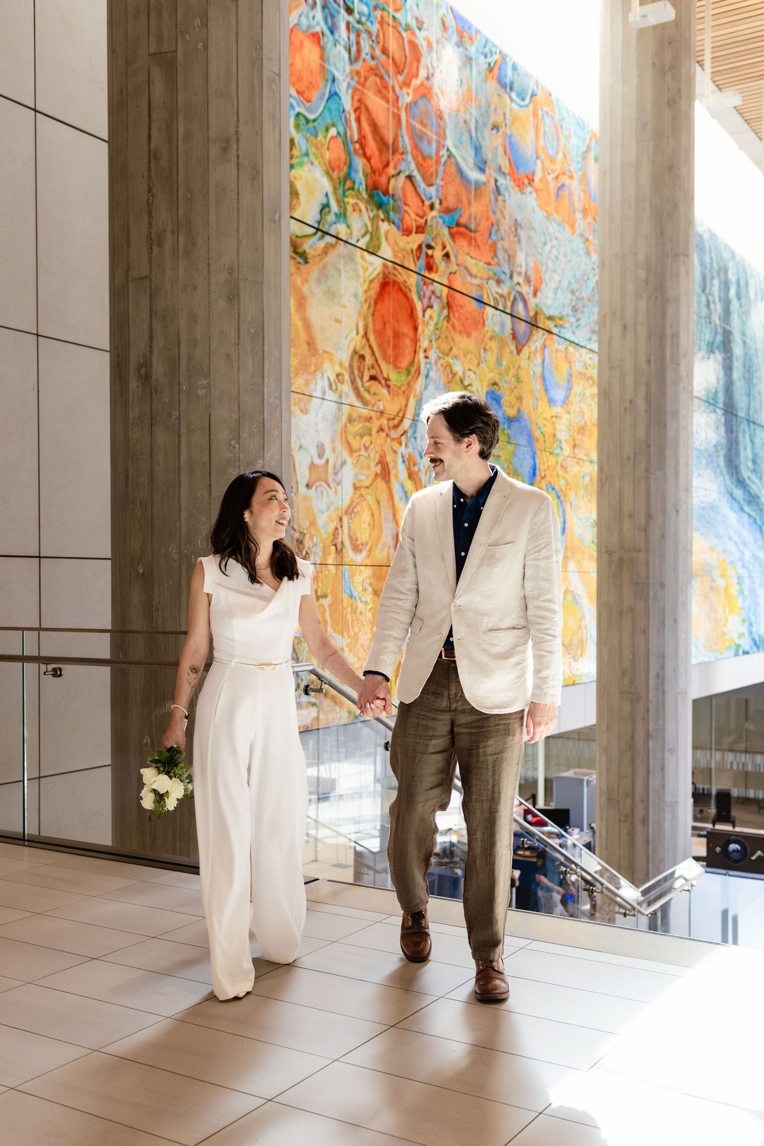 Bride and groom walk hand in hand inside a modern building with a vibrant colorful mural backdrop.