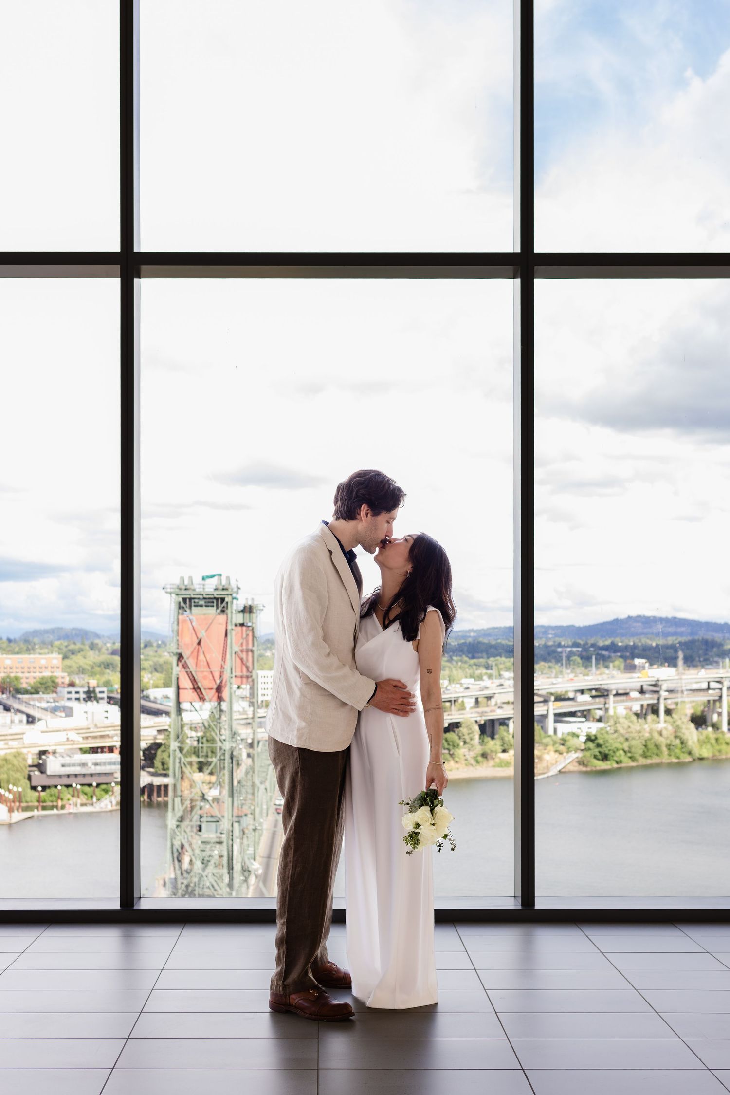 Couple sharing a romantic kiss in wedding attire before large windows with a cityscape and river view.