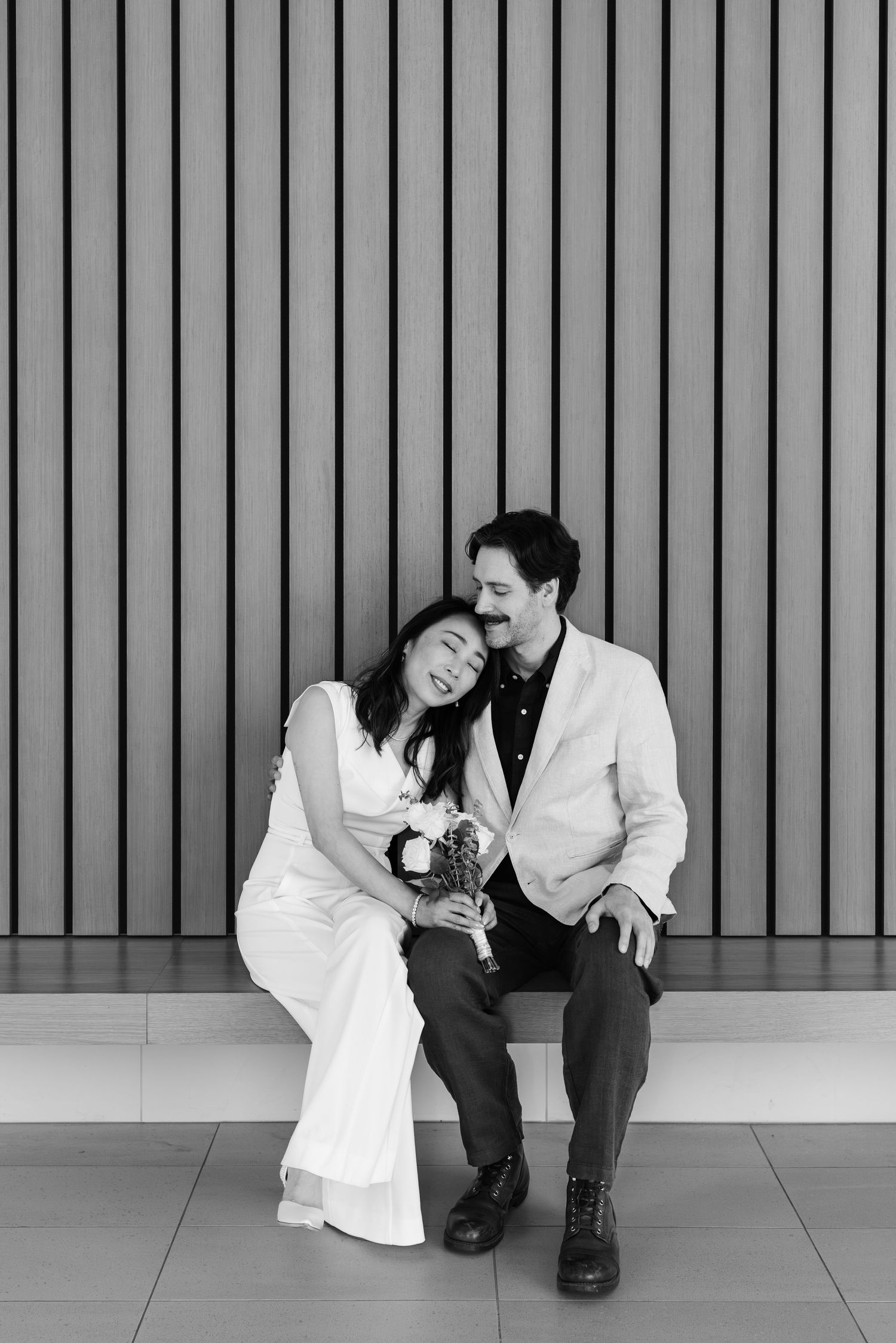Black and white photo of a couple in wedding attire sitting together, smiling and leaning on each other against a slatted wall.