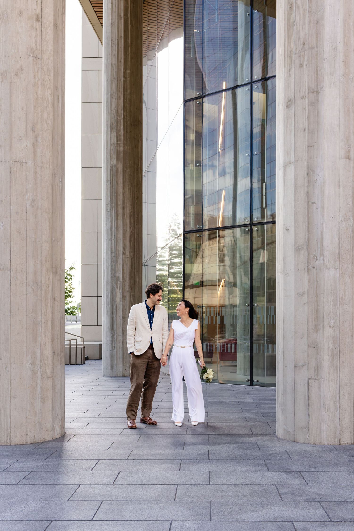 Couple in elegant attire walking between tall columns outside a modern glass building.