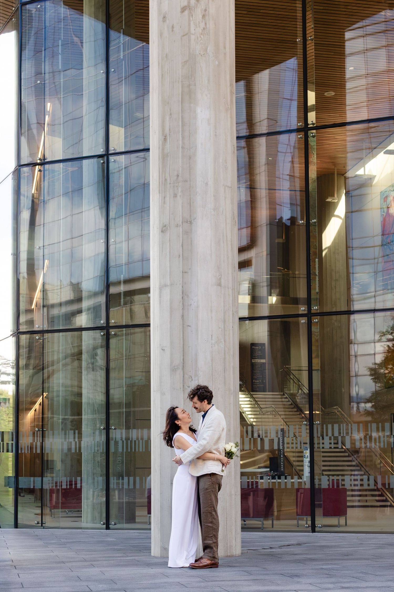 Bride and groom embracing near a modern glass building column on their wedding day.