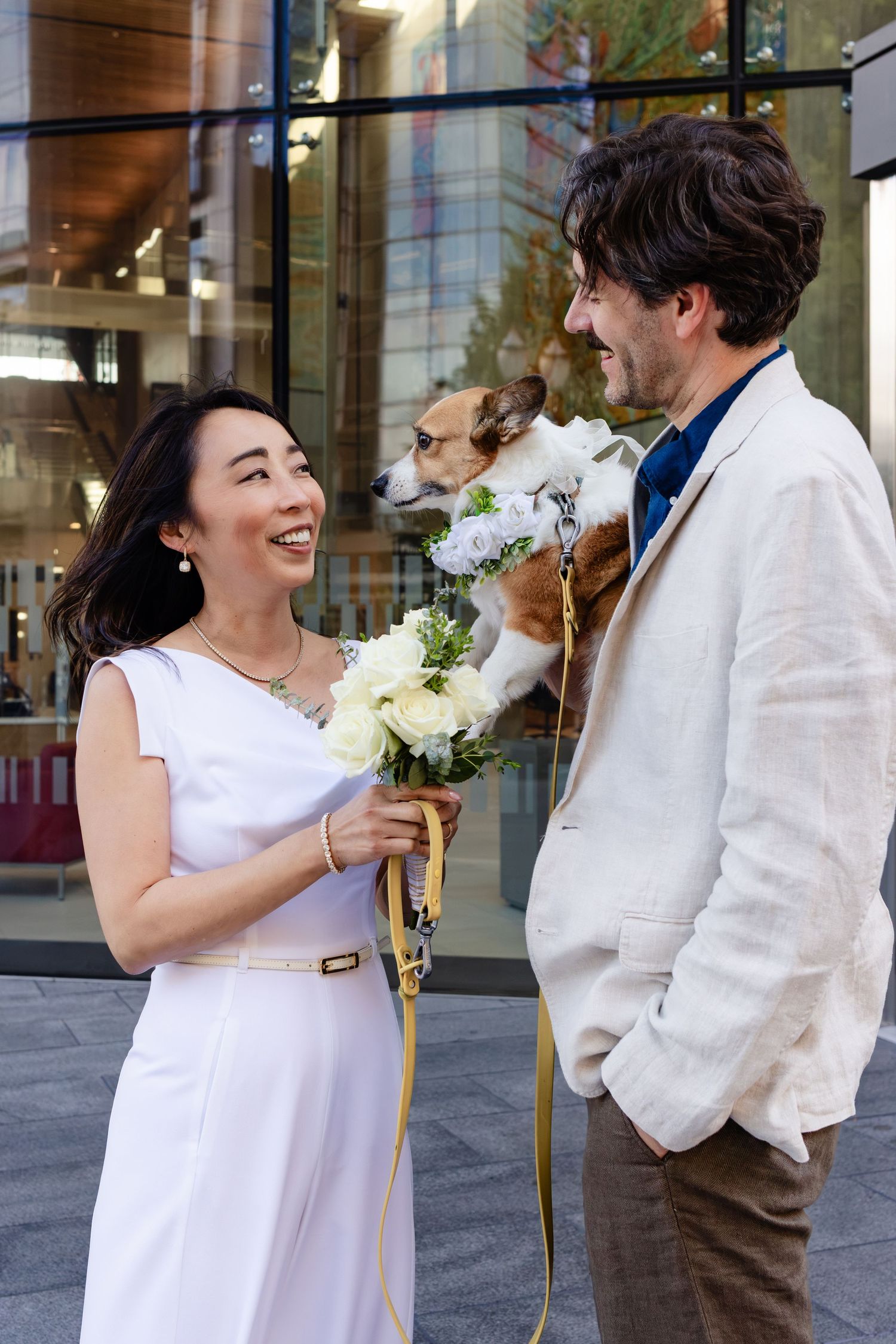 Smiling bride in white dress holds bouquet beside groom in linen suit with dog on leash outside modern building.