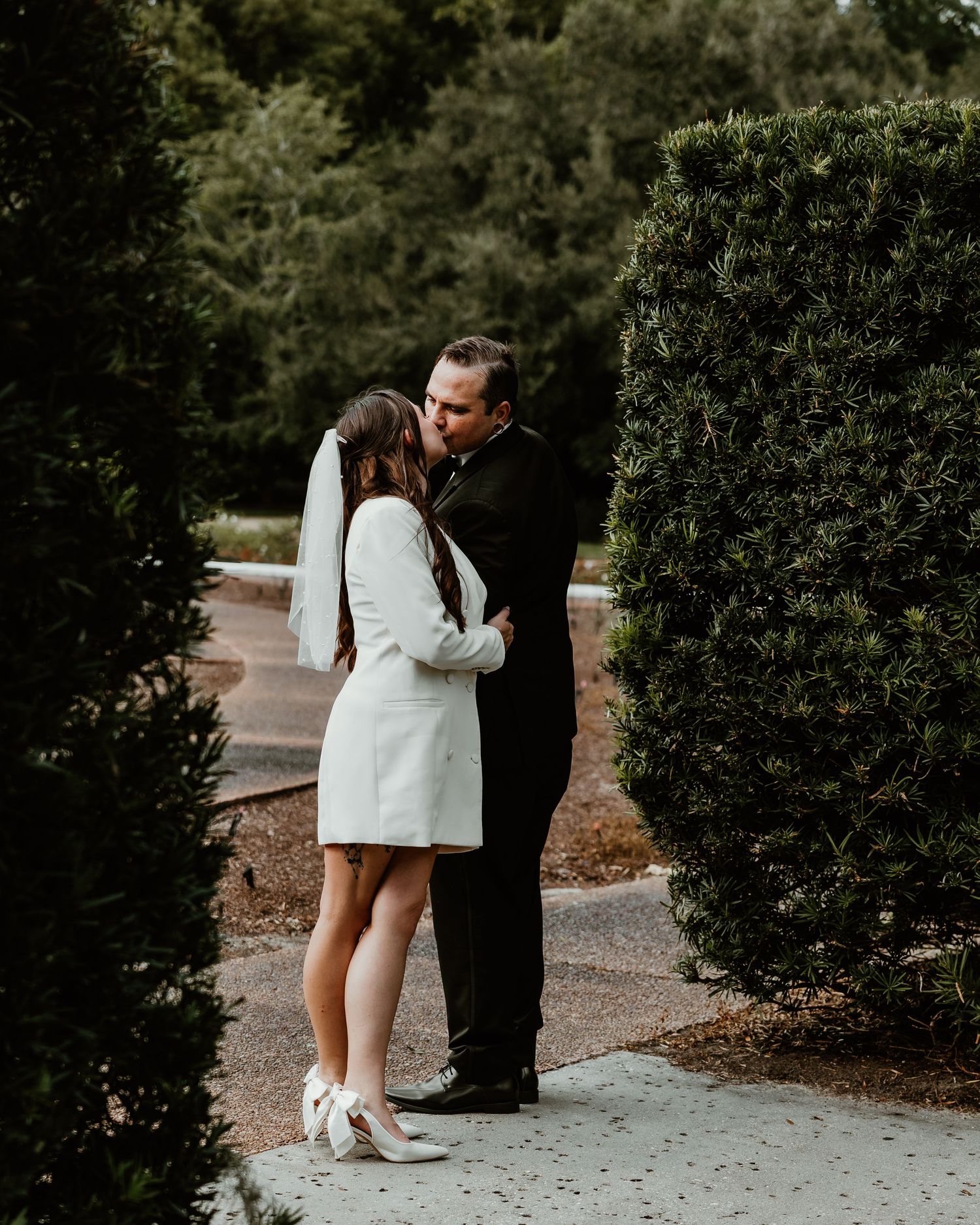 A bride in a white blazer dress and veil shares a romantic kiss with her groom in a black suit among lush green hedges.