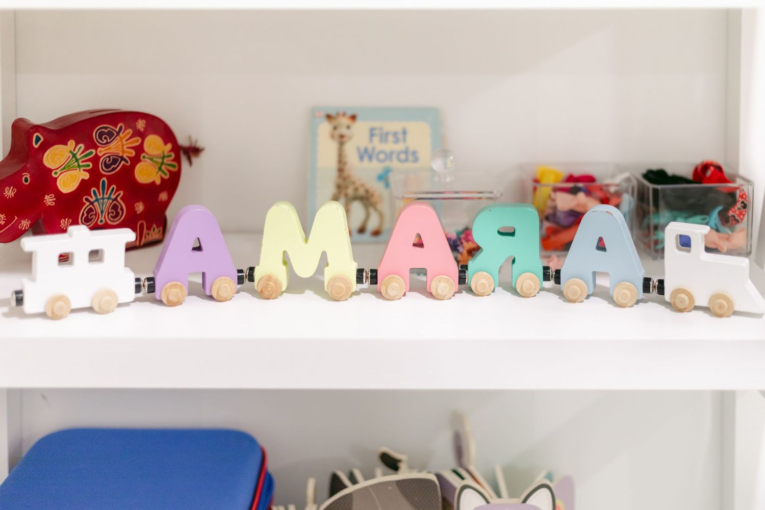 Colorful wooden letter train spelling AMARA on a white shelf with children's books and toys in background.
