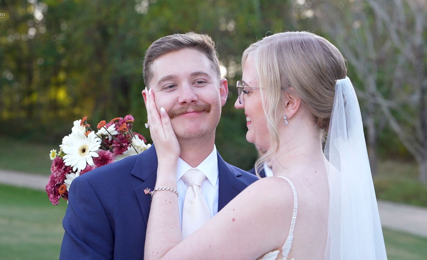 Bride in white veil touches groom's face tenderly outdoors, groom wearing blue suit holds bouquet at wedding.