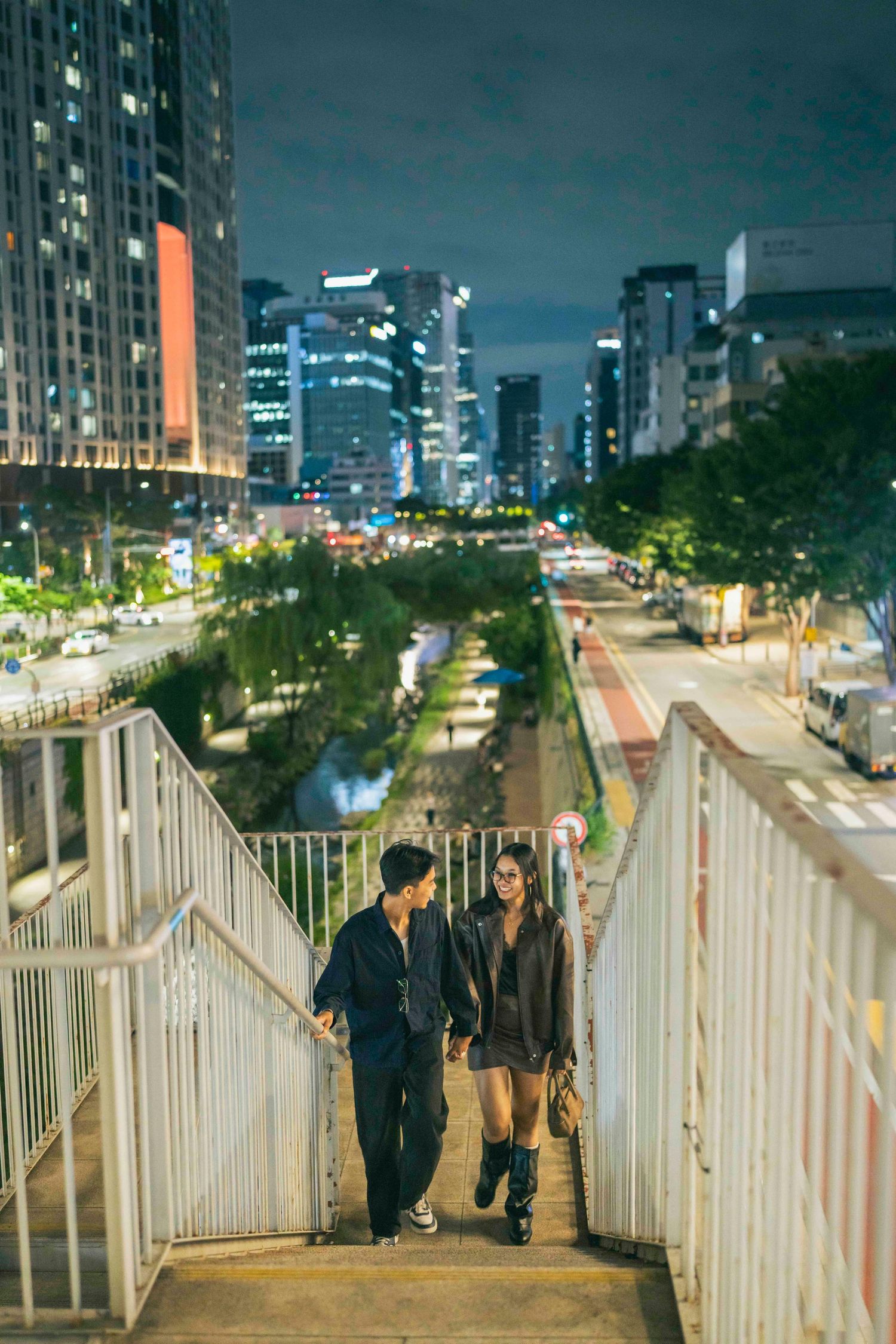 Couple walking up city stairs at night with illuminated skyscrapers and busy street in background.