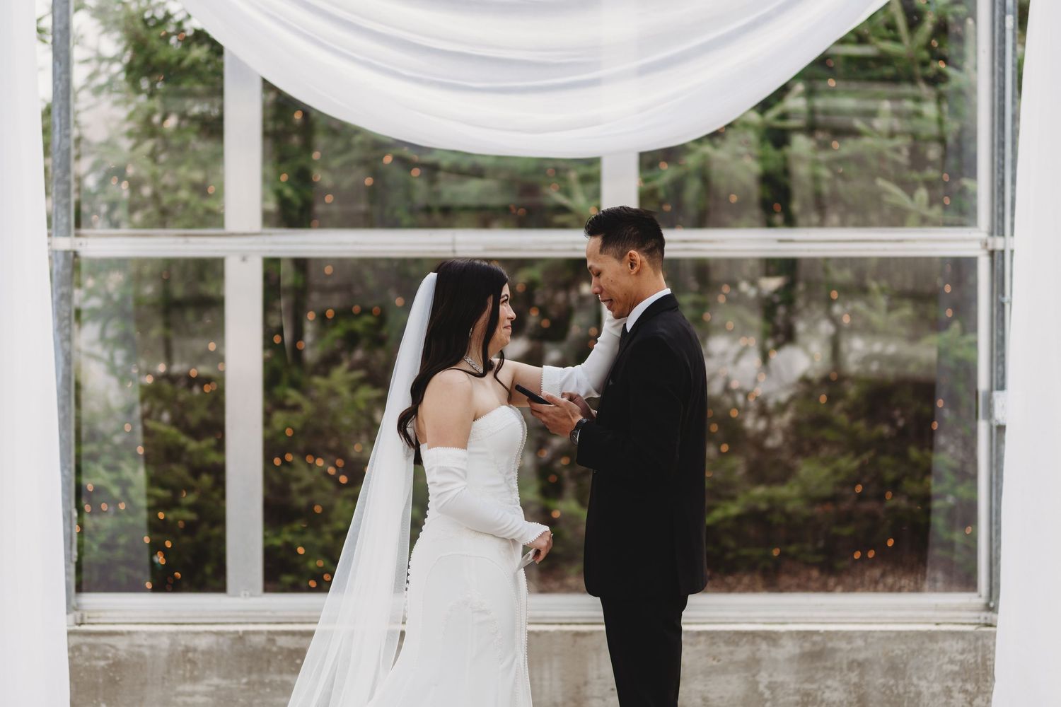 Bride and groom exchange vows at elegant indoor wedding ceremony with white draping and large windows.
