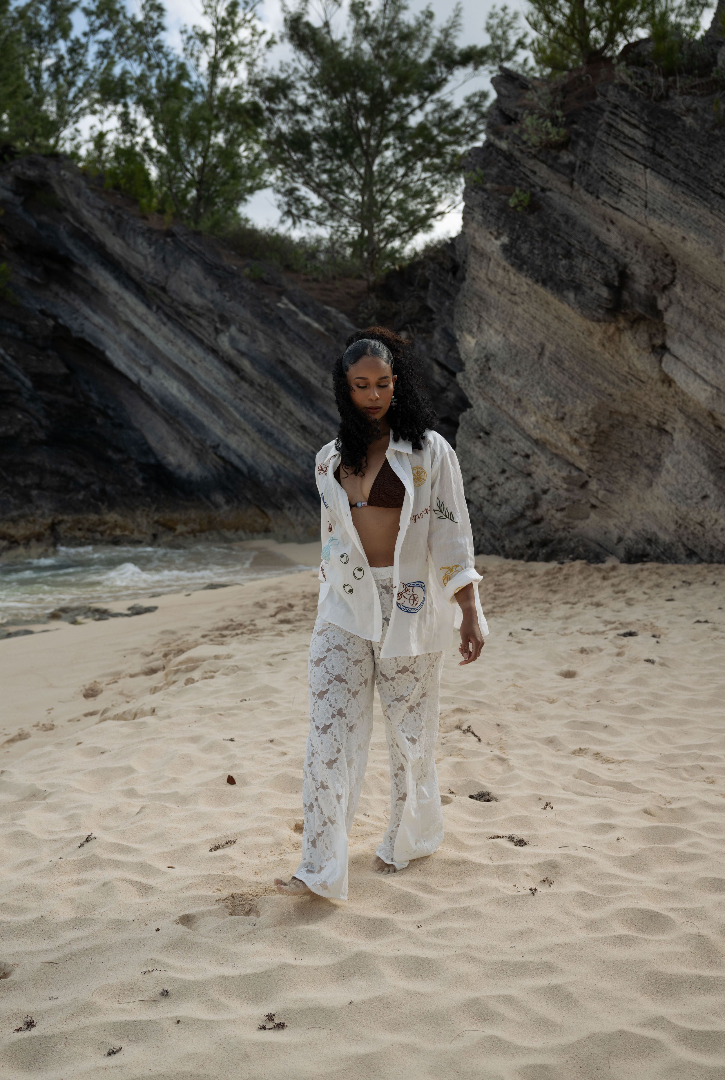 Woman in white linen outfit walking on sandy beach with rocky cliffs and green trees in background.