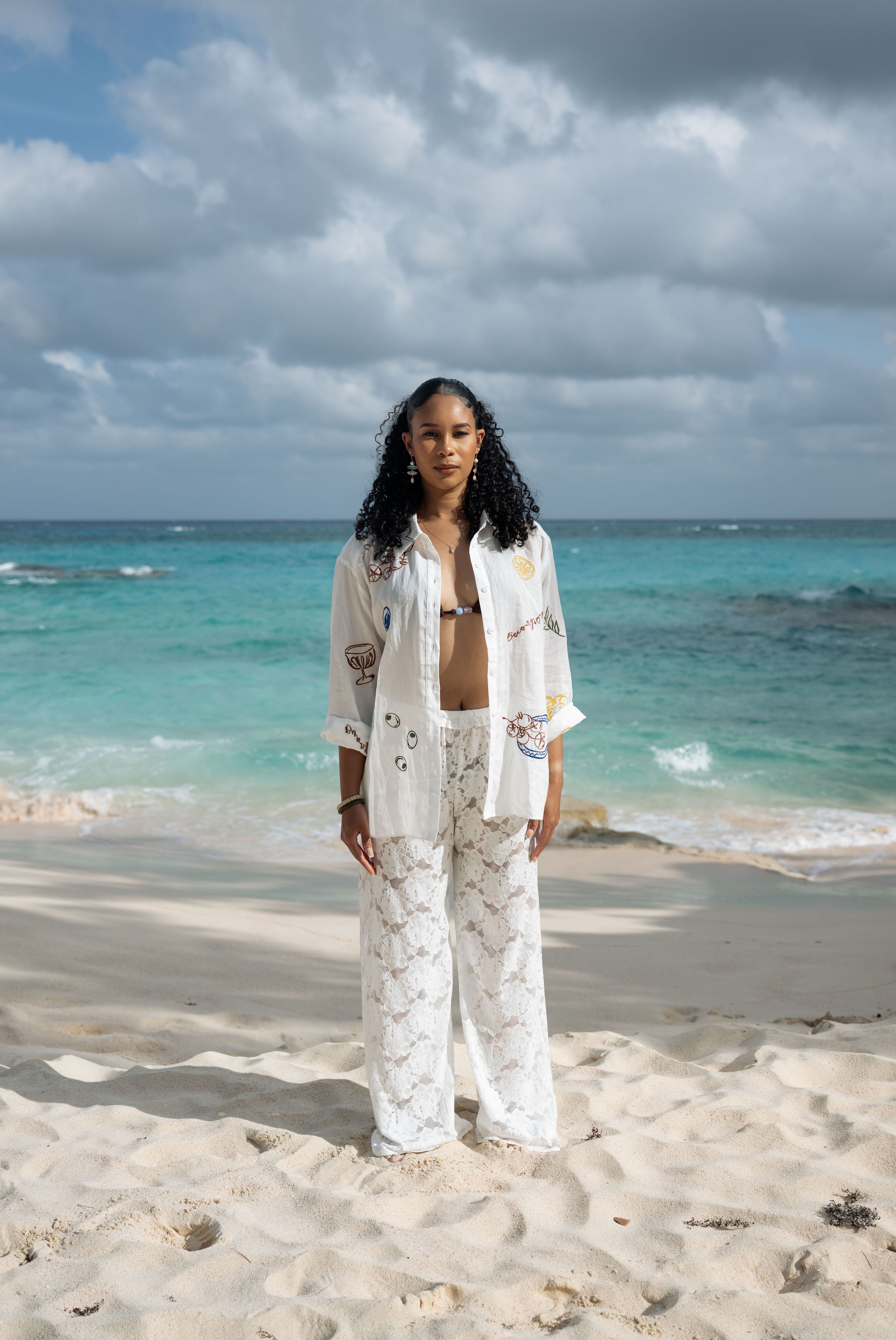 Woman in white lace pants and embroidered shirt stands on a sandy beach with turquoise ocean backdrop.