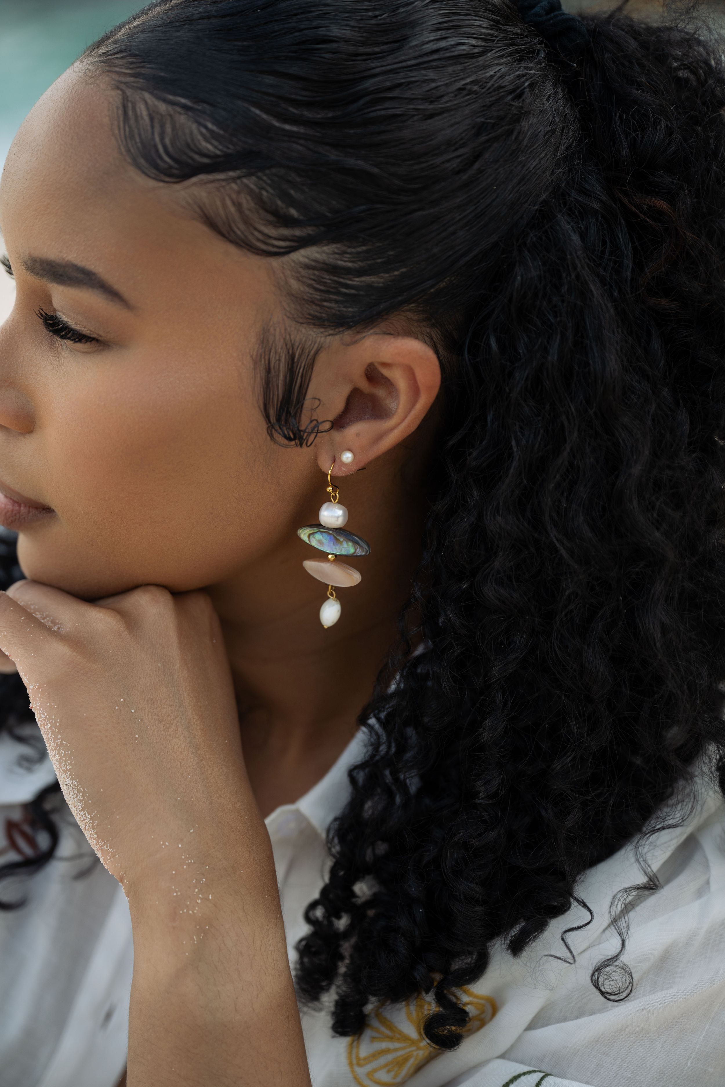 Woman wearing elegant gold drop earrings with turquoise stones and pearl accents, resting chin on hand.