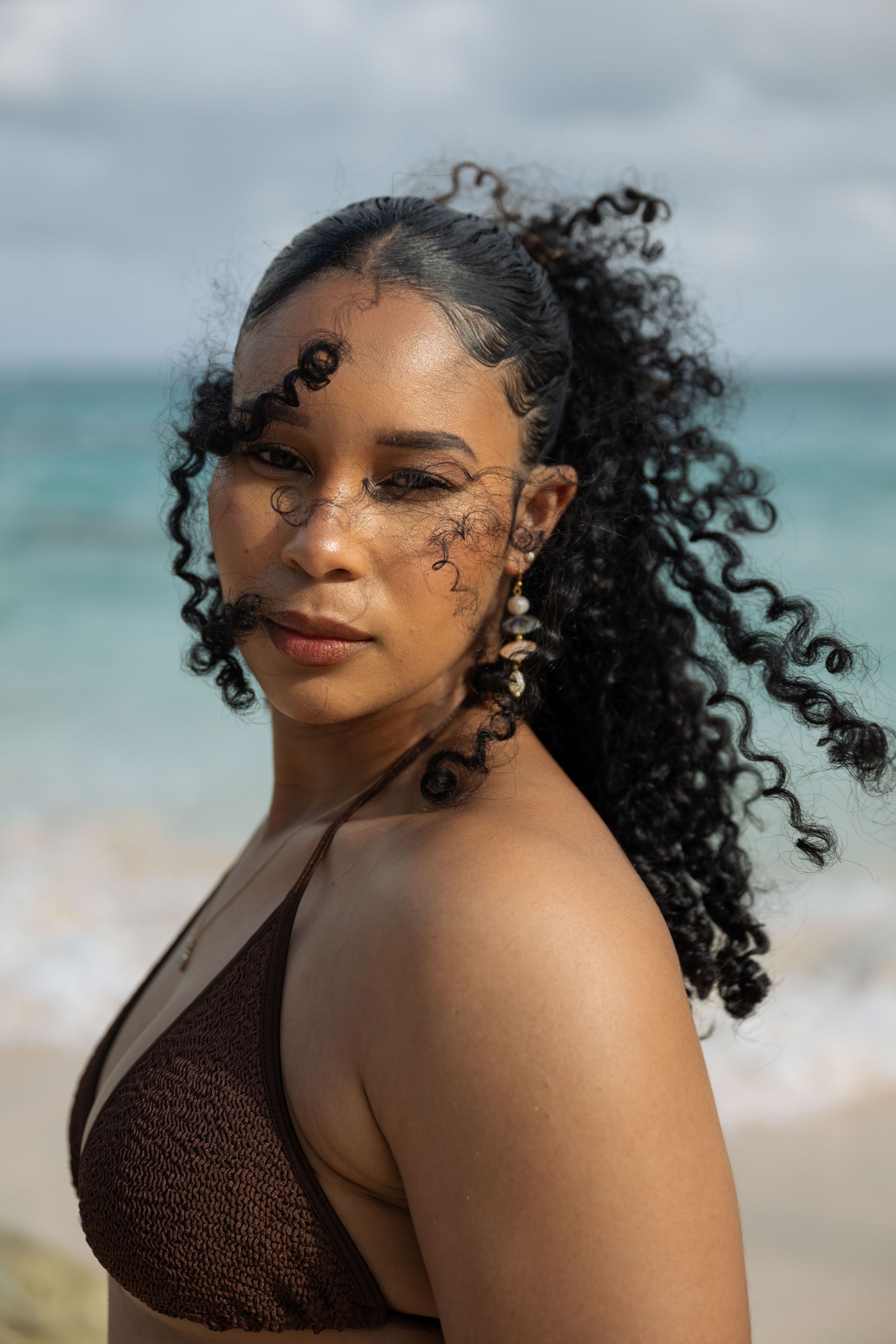 Woman with curly hair and face art posing on a beach in a brown bikini top with ocean waves behind her.