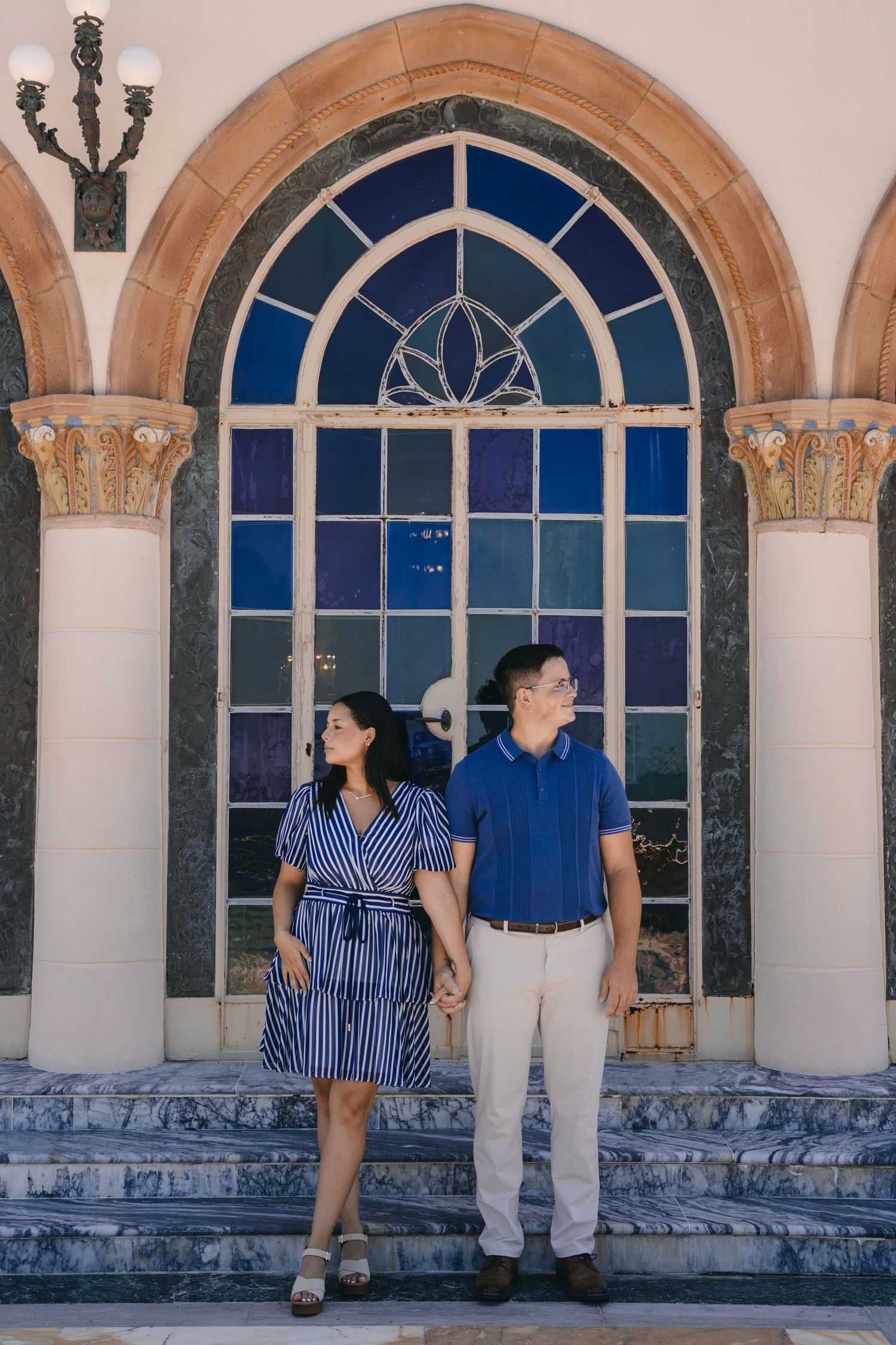 Couple holding hands standing on steps before ornate arched window with stained glass, woman in blue dress, man in blue shirt.