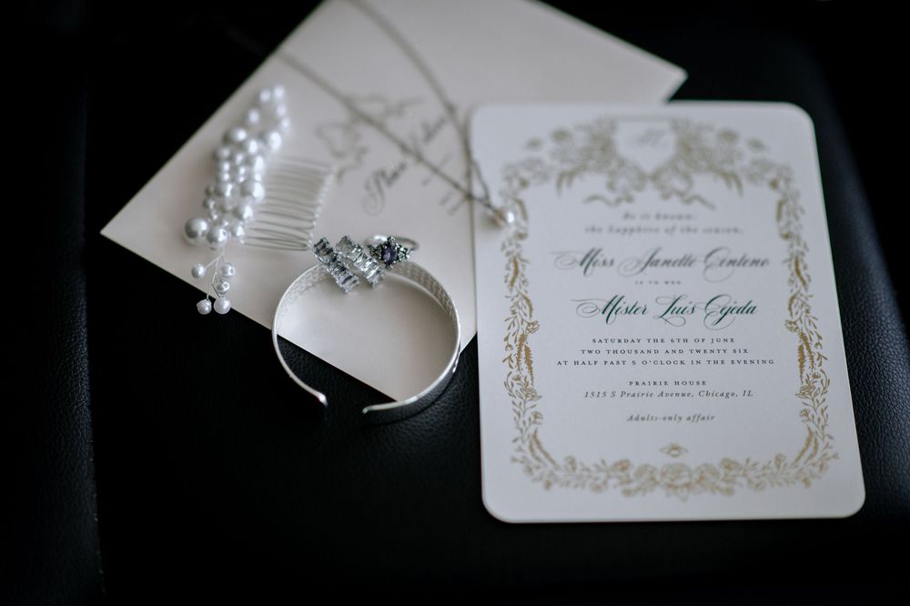 Elegant wedding invitation with gold border beside pearl jewelry, diamond ring, and hair comb on dark surface.