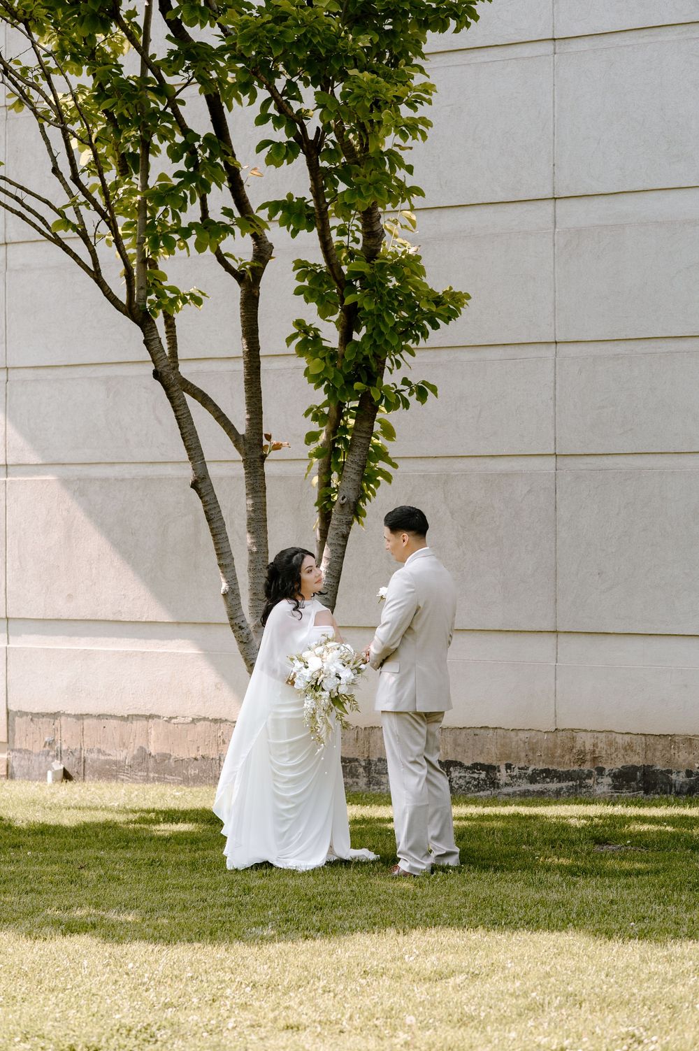 Bride and groom stand facing each other on green lawn beside tree against modern stone wall in wedding attire.