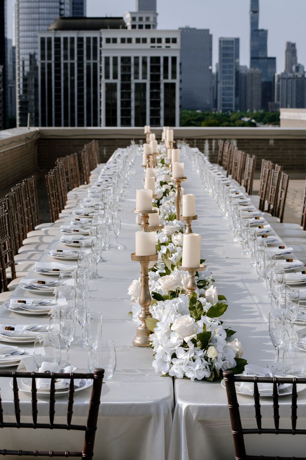 Elegant rooftop wedding reception with long white tables, floral centerpieces, candelabras, and city skyline views.
