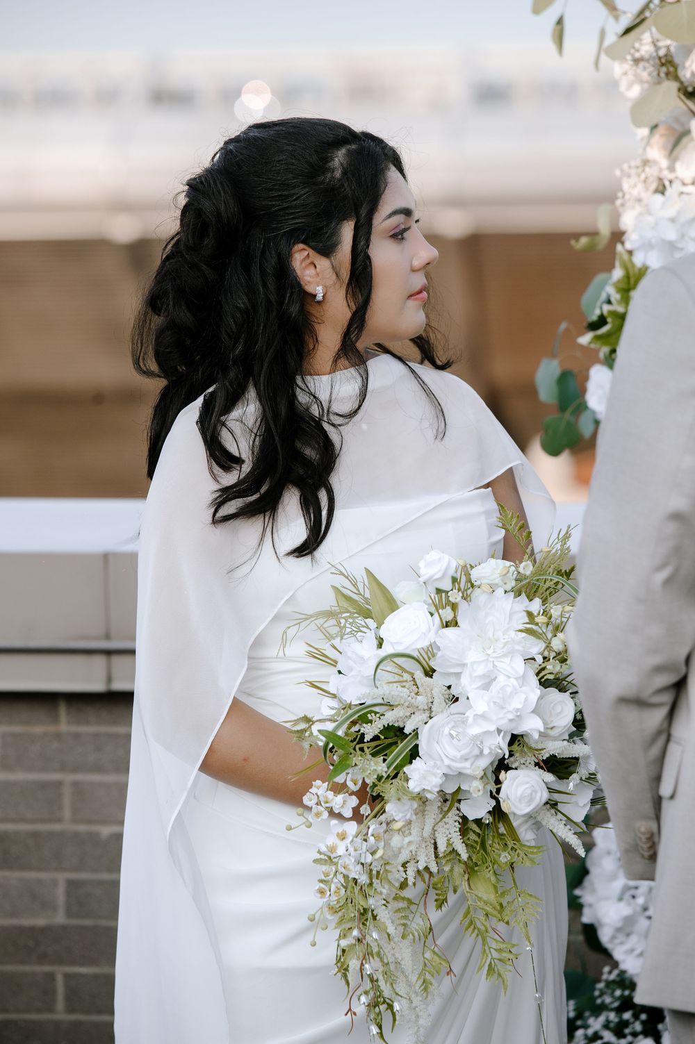 Bride with dark hair holds white floral bouquet wearing elegant white cape wedding gown at outdoor ceremony.
