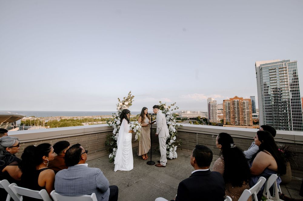 Rooftop wedding ceremony with couple at floral arch, guests seated in white chairs, ocean and city skyline backdrop.