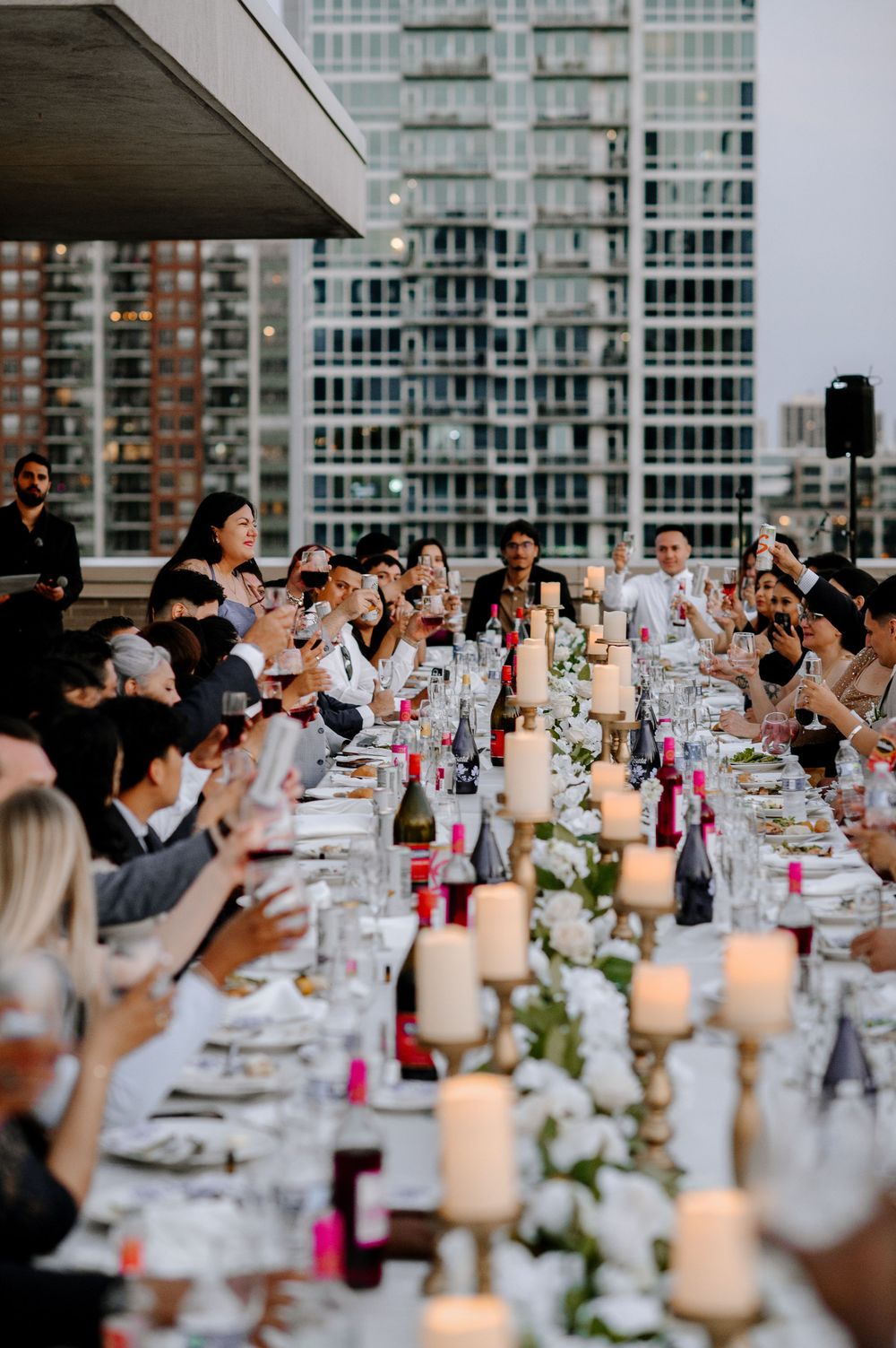 Guests raise glasses in a toast at an elegant rooftop dinner party with candles and city skyline backdrop.