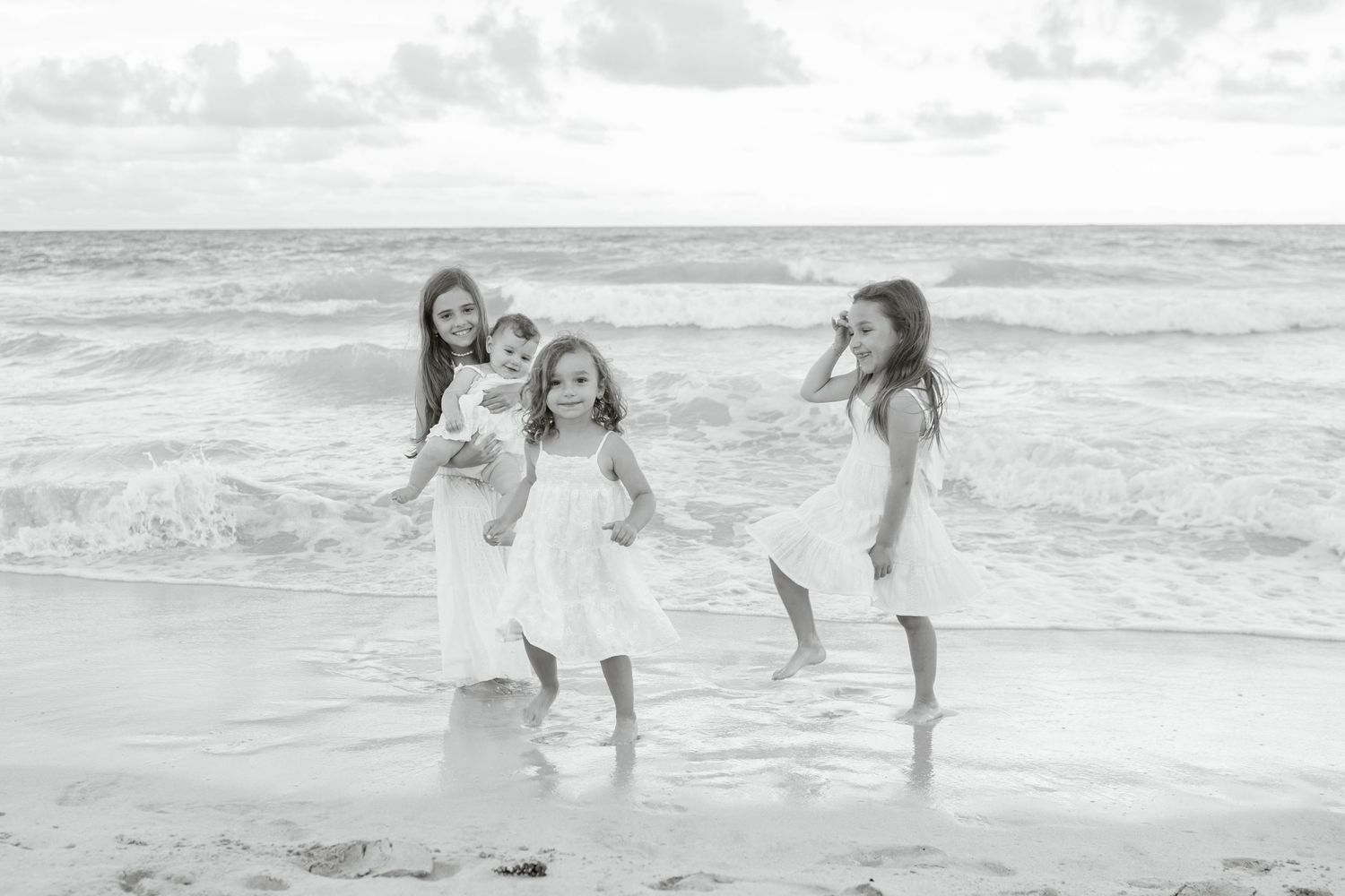 Black and white photo of four girls in white dresses playing on Kailua beach with ocean waves behind them.