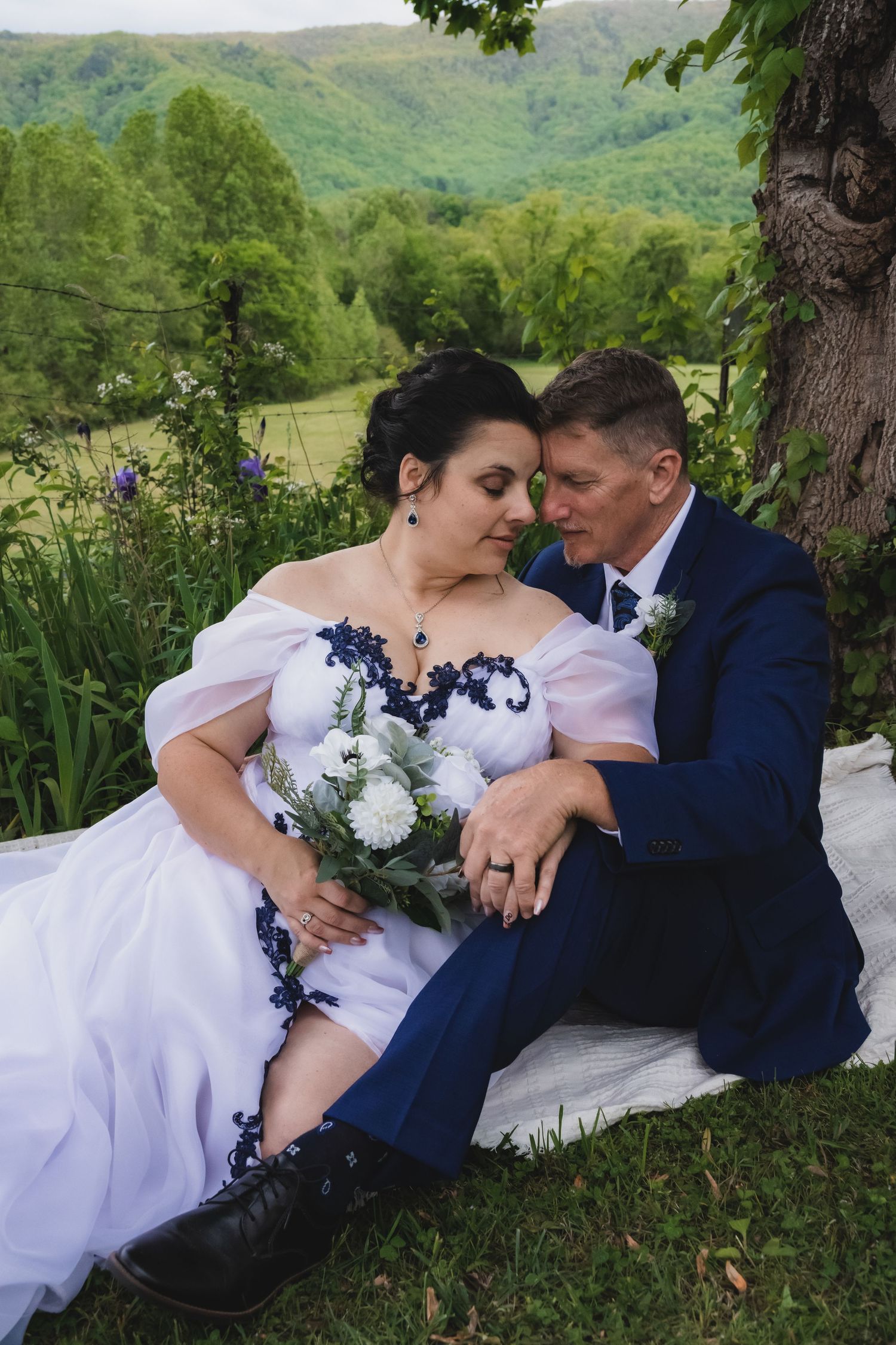 Bride and groom embrace in tall grass field with dramatic purple sunset sky and mountain forest backdrop.