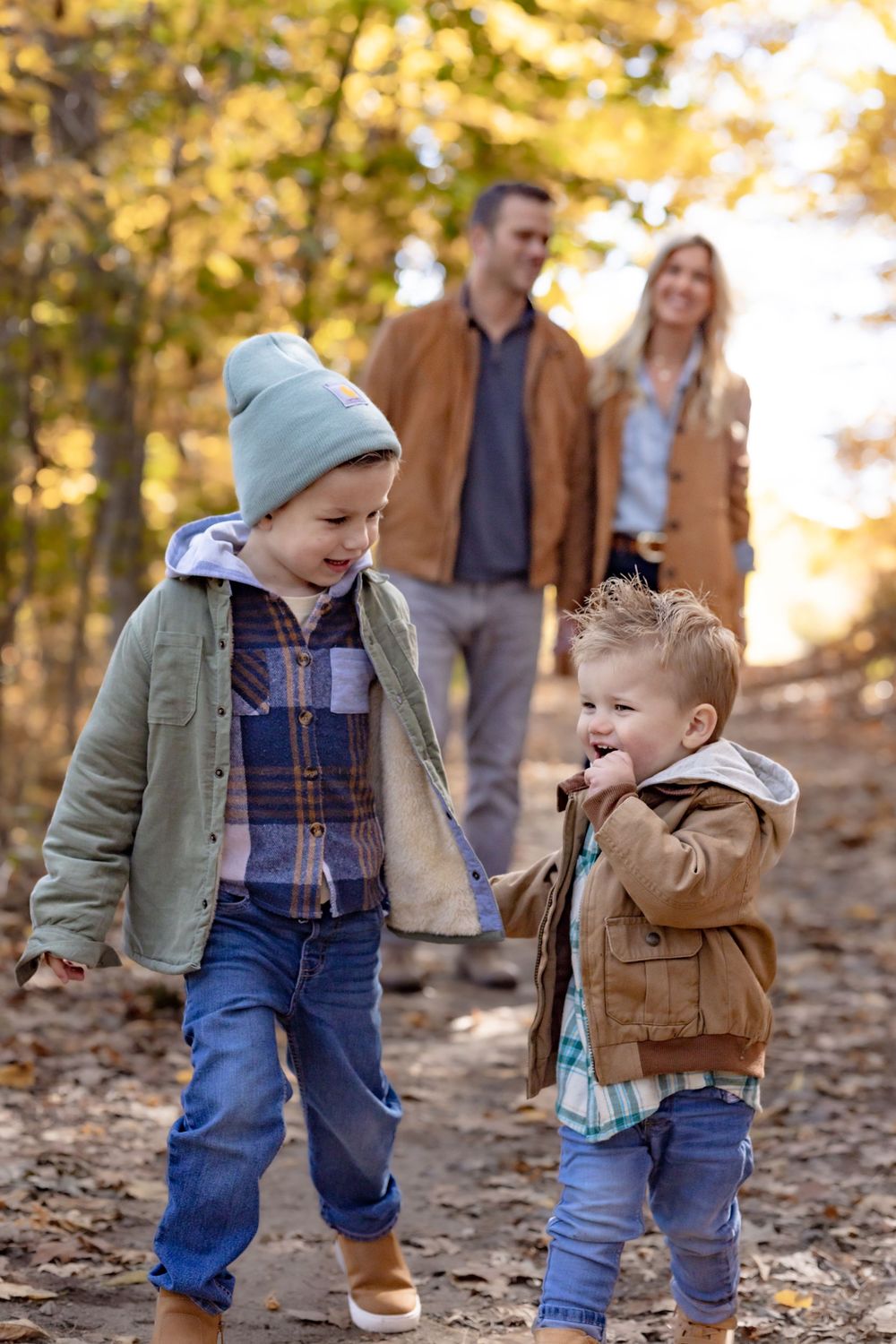 Two young boys laughing and playing on an autumn forest path with their parents walking behind them.