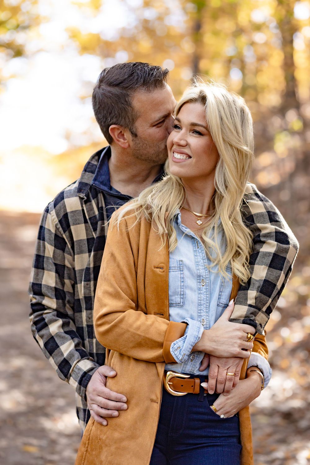 Couple embracing outdoors in autumn, woman smiling in tan jacket, man kissing her cheek, fall foliage background.