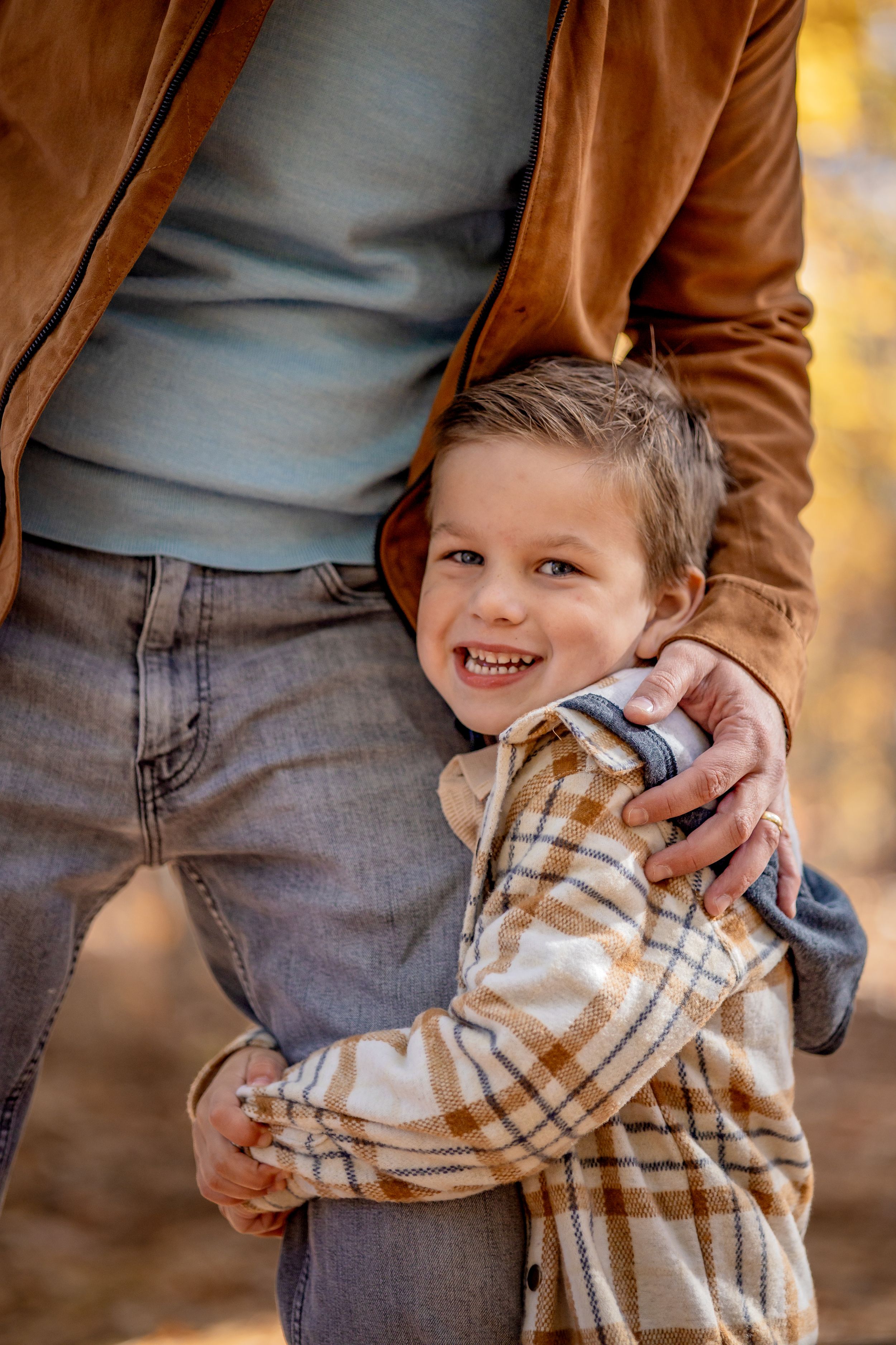 A smiling young boy hugs an adult's leg outdoors in autumn, wearing a plaid shirt with warm fall colors in the background.