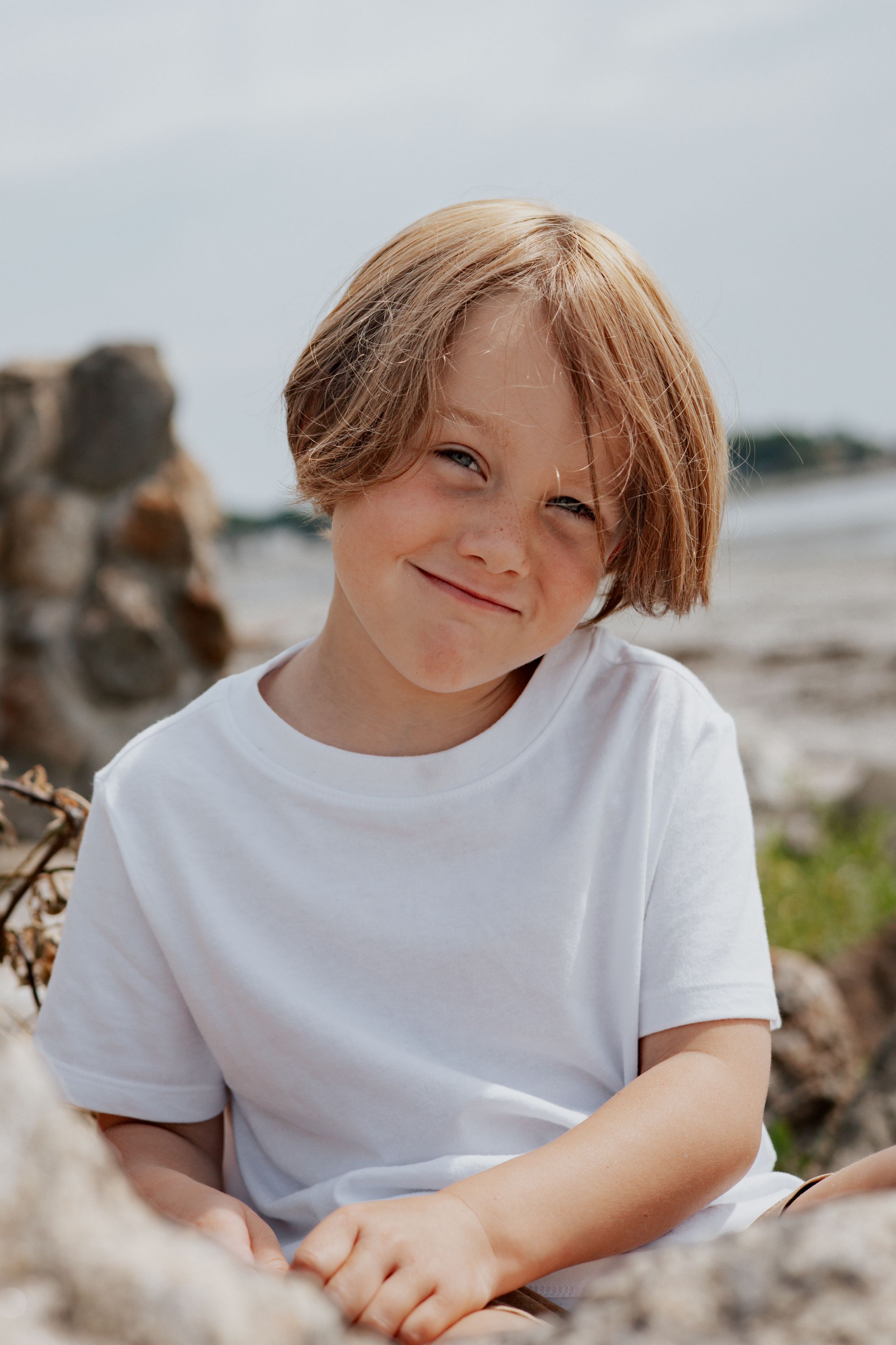 Smiling blond boy in white t-shirt sitting on rocks at the beach on a sunny day.