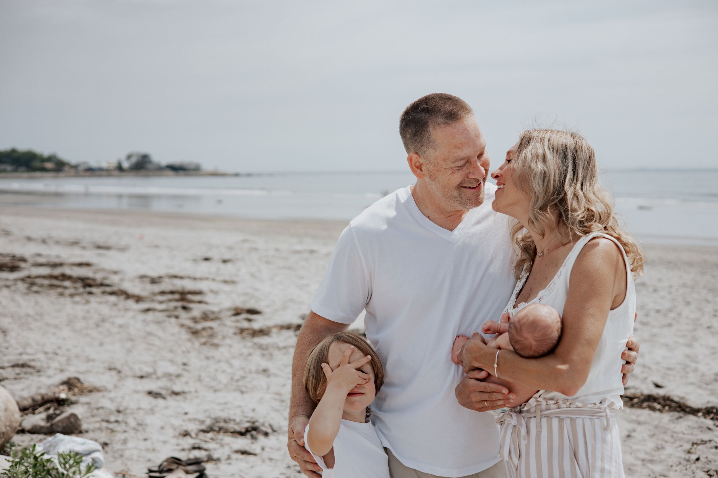 A happy couple with two children share a tender moment on a sandy beach, with calm ocean waters in the background.