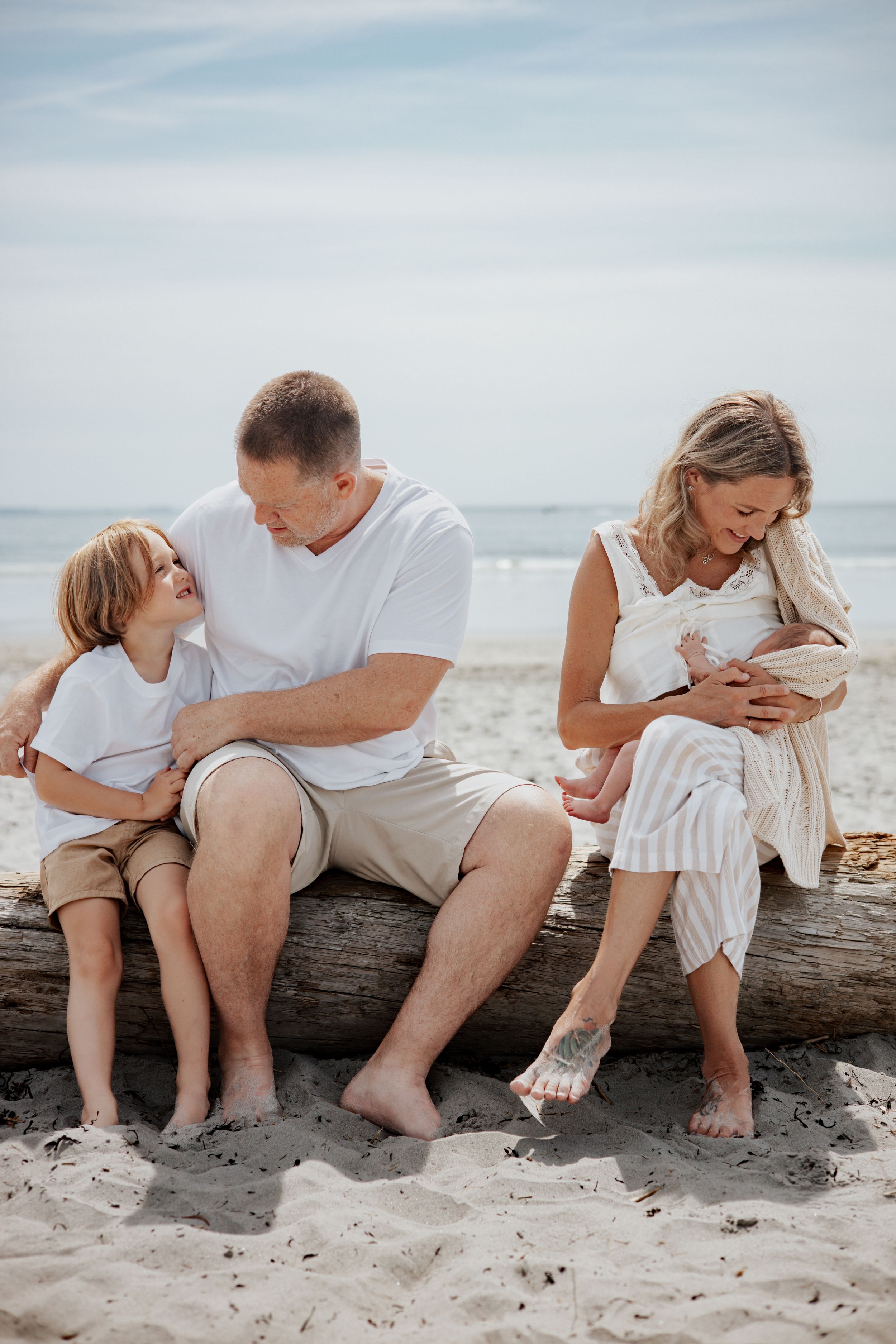 Family sitting on driftwood at beach, parents with young boy and newborn baby in natural light.