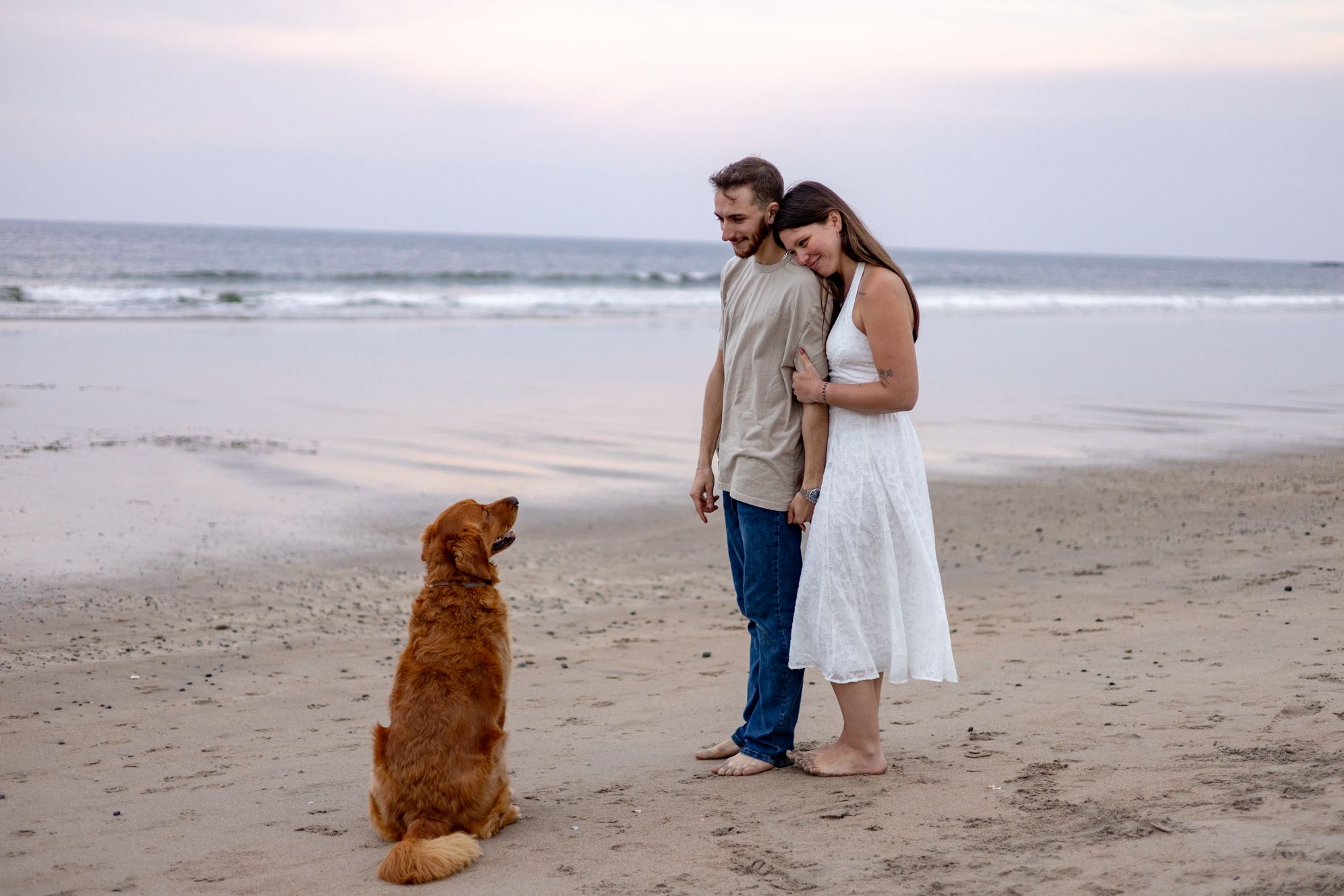 Couple in romantic embrace on sandy beach at dusk with golden retriever dog sitting nearby.