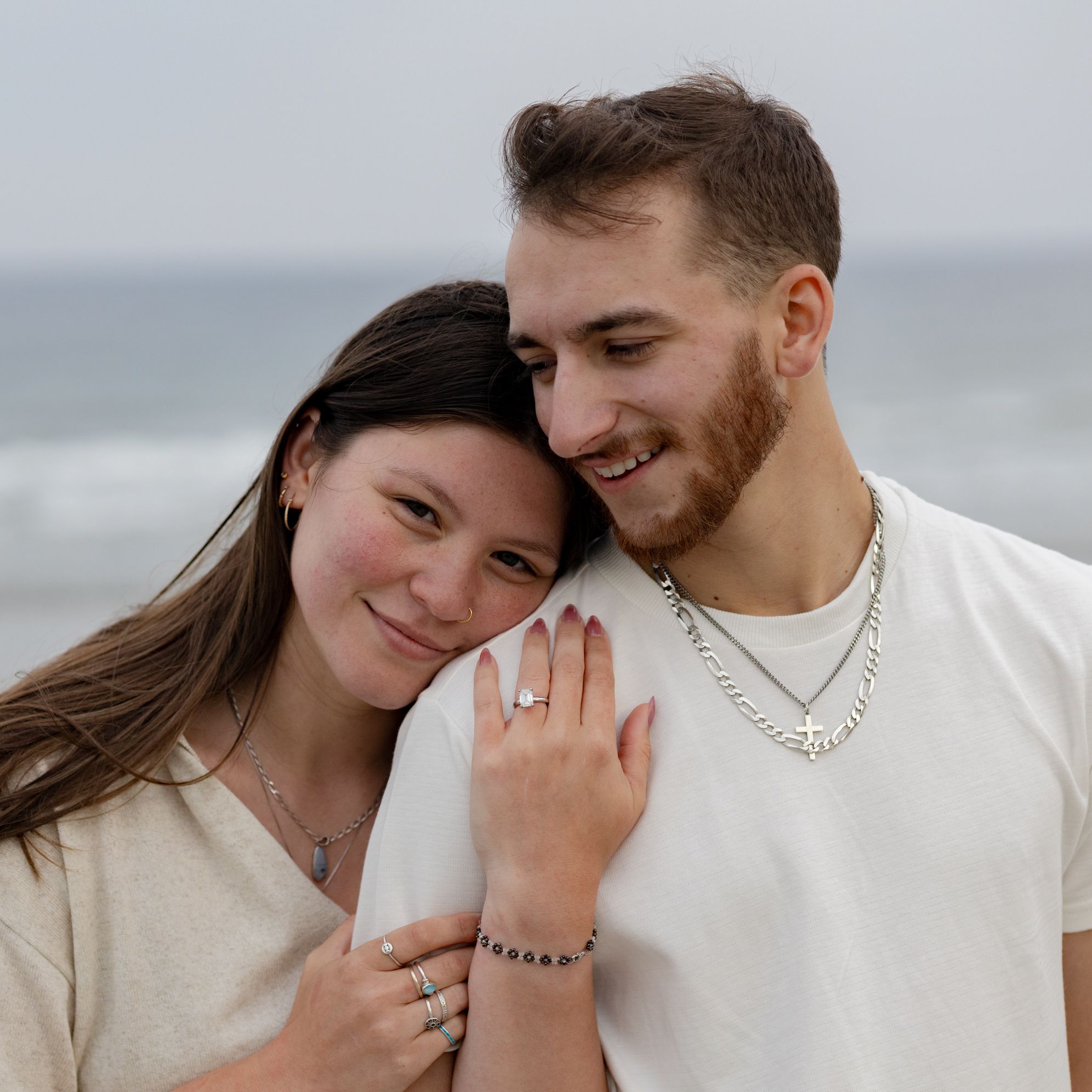 Couple embracing on beach, woman showing engagement ring, both wearing silver jewelry, ocean in background.