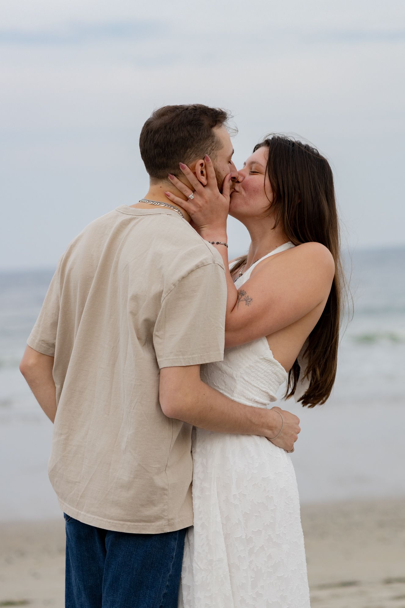 A couple sharing a romantic kiss on the beach, woman in white dress and man in beige shirt with blue jeans.