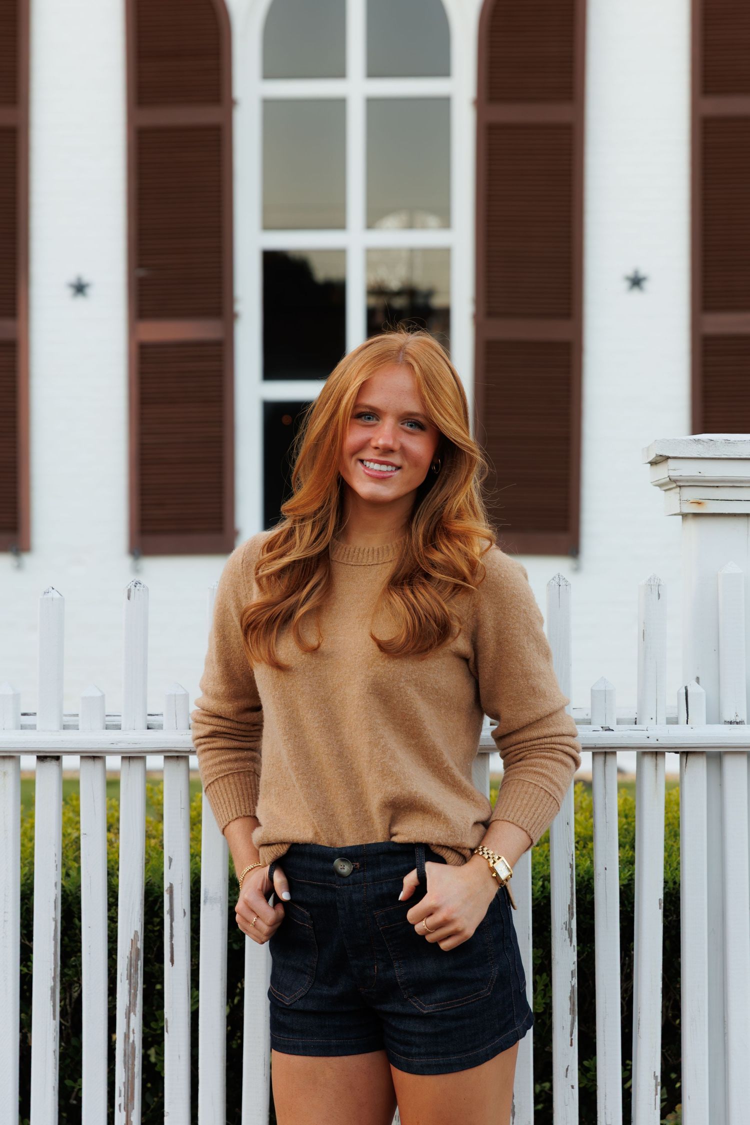 Smiling redhead woman in tan sweater and black denim shorts standing by white picket fence.