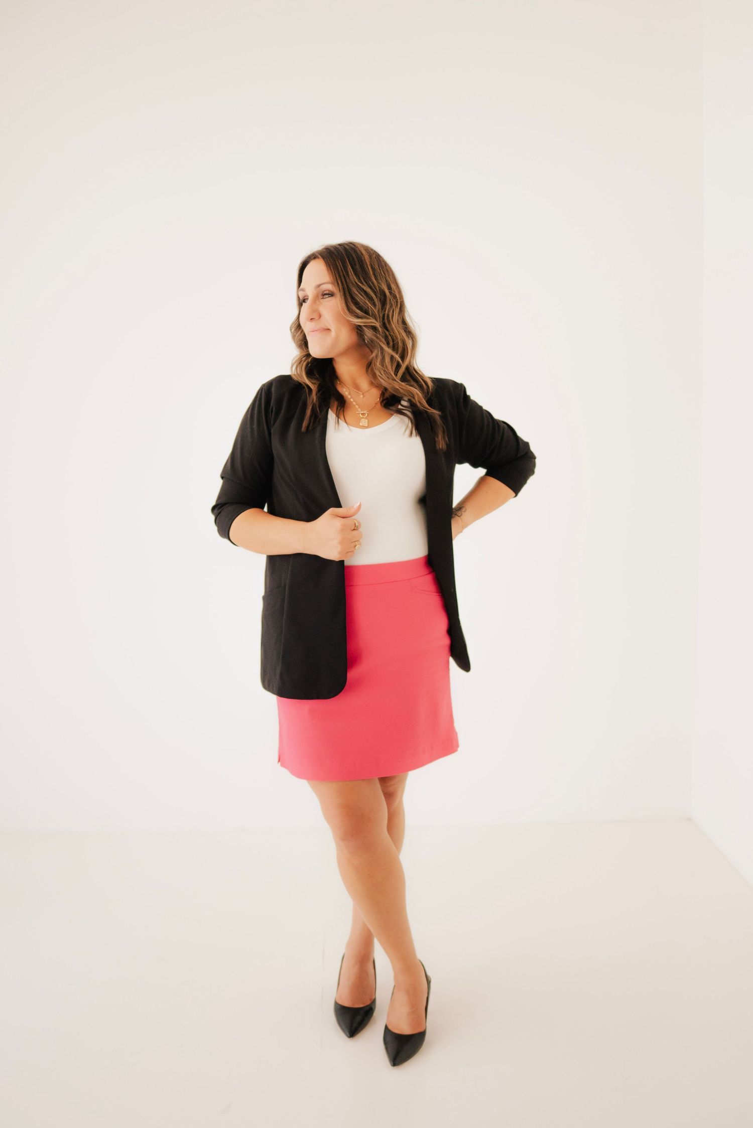 Woman in pink mini skirt, white top, black blazer and heels poses confidently against white background.