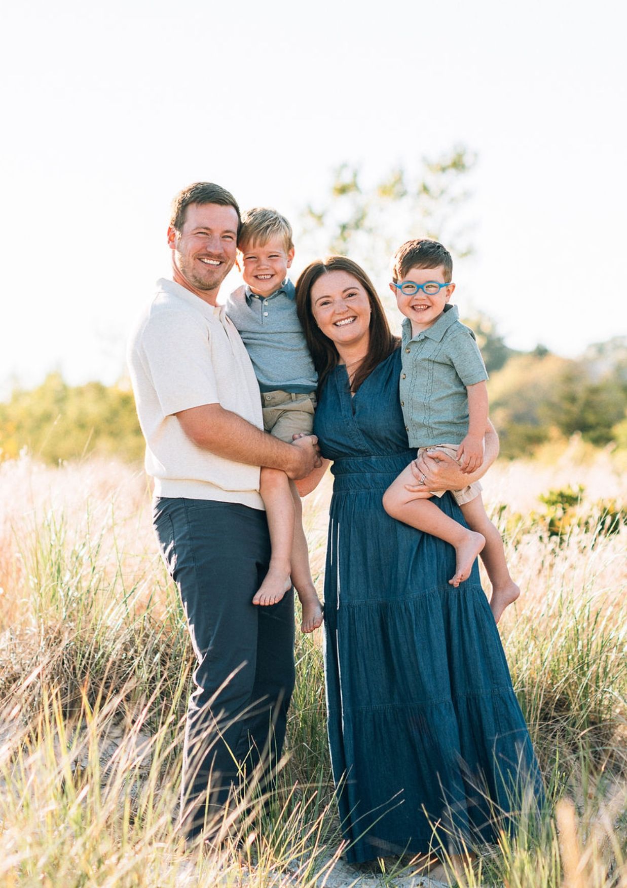 Happy family of four posing outdoors in a sunny field, parents holding two young boys, wearing casual summer outfits.